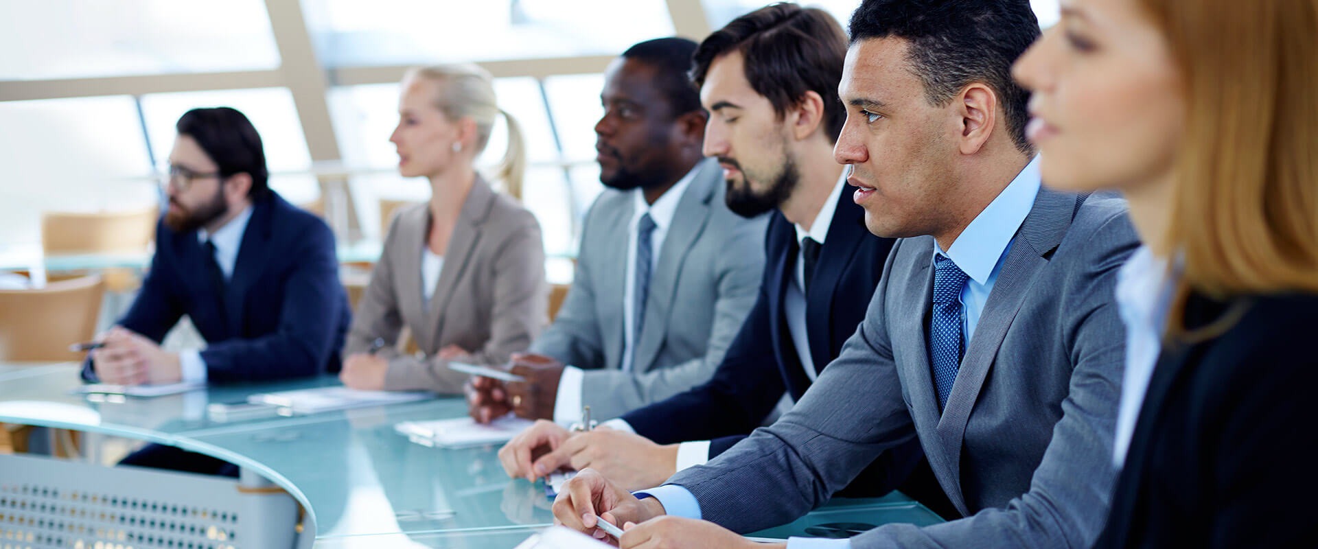 Grupa profesjonalistów w garniturach uczestniczy w spotkaniu przy szklanym stole, z widocznymi notatkami i długopisami, w jasnym pomieszczeniu biurowym.