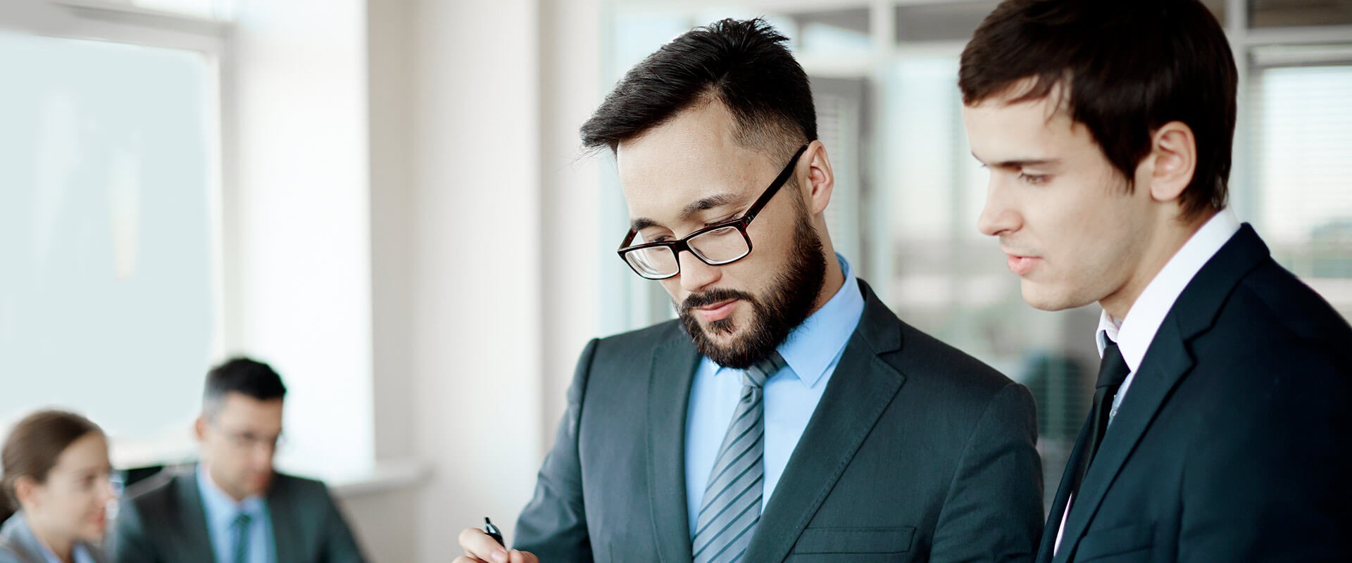 Dwóch eleganckich mężczyzn w garniturach analizuje dokument, jeden z nich trzyma długopis, w tle rozmyte postacie przy oknie.