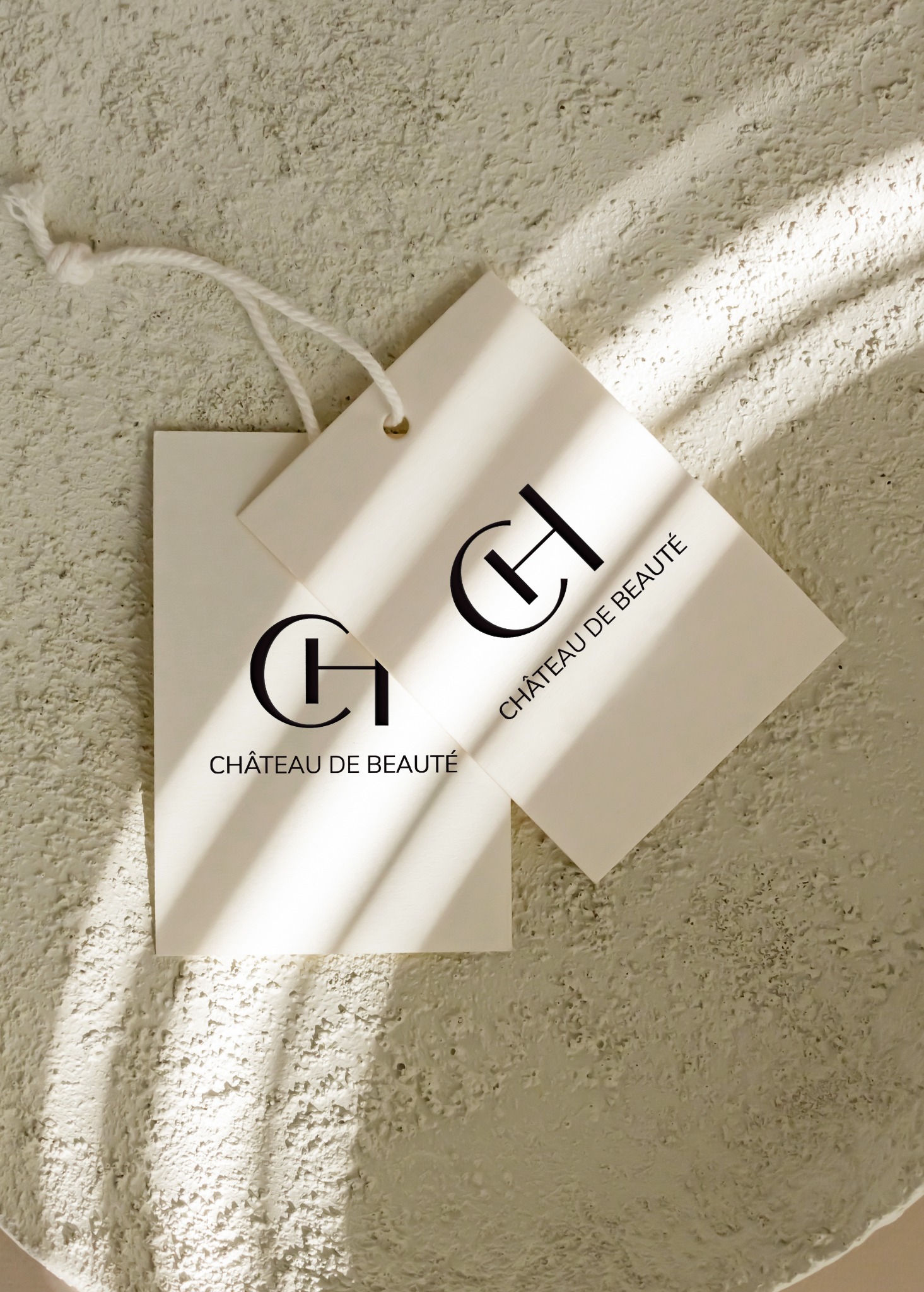 Dwie kwadratowe etykiety z minimalistycznym czarnym logo 'CH' i napisem 'Château de Beauté' leżą na teksturowanym, beżowym tle w naturalnym świetle.
