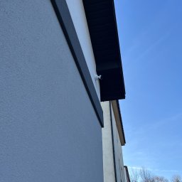 BARWAS SP. Z O. O. - Kamera monitoringu zamontowana na elewacji budynku, widoczna z dołu pod kątem. Biała kamera na tle błękitnego nieba i fragmentu ściany budynku.