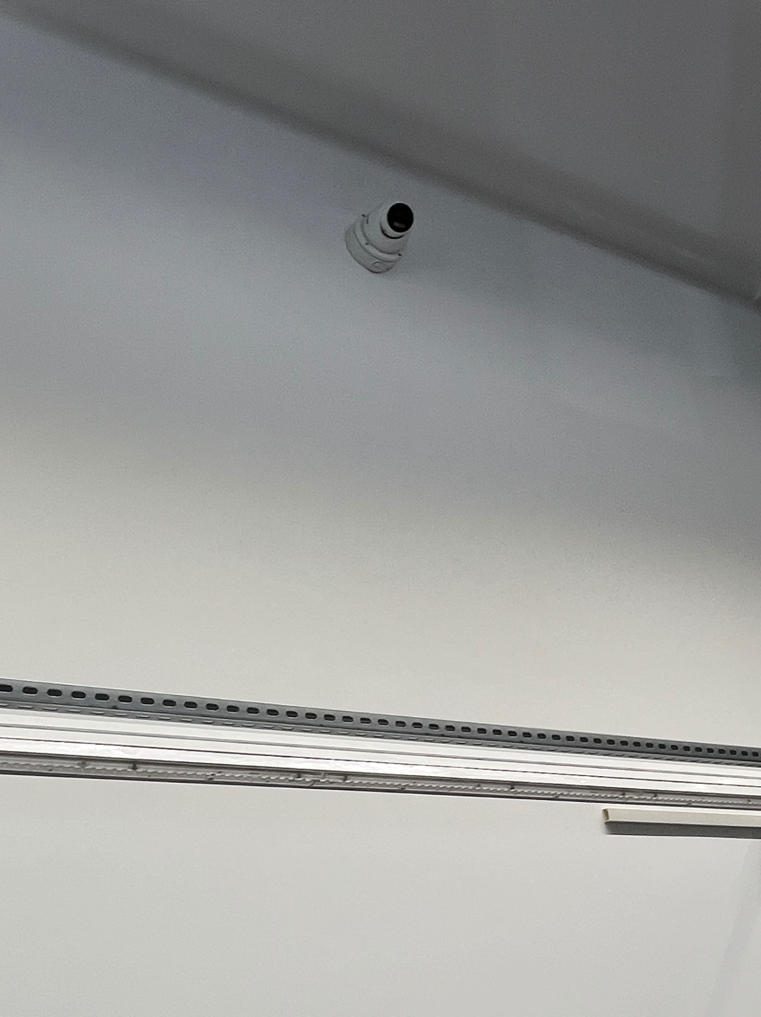 Kamera monitoringu kopułkowa zamontowana na suficie, widoczna z dołu. Szara listwa montażowa wzdłuż ściany. Białe tło sufitu i ściany.