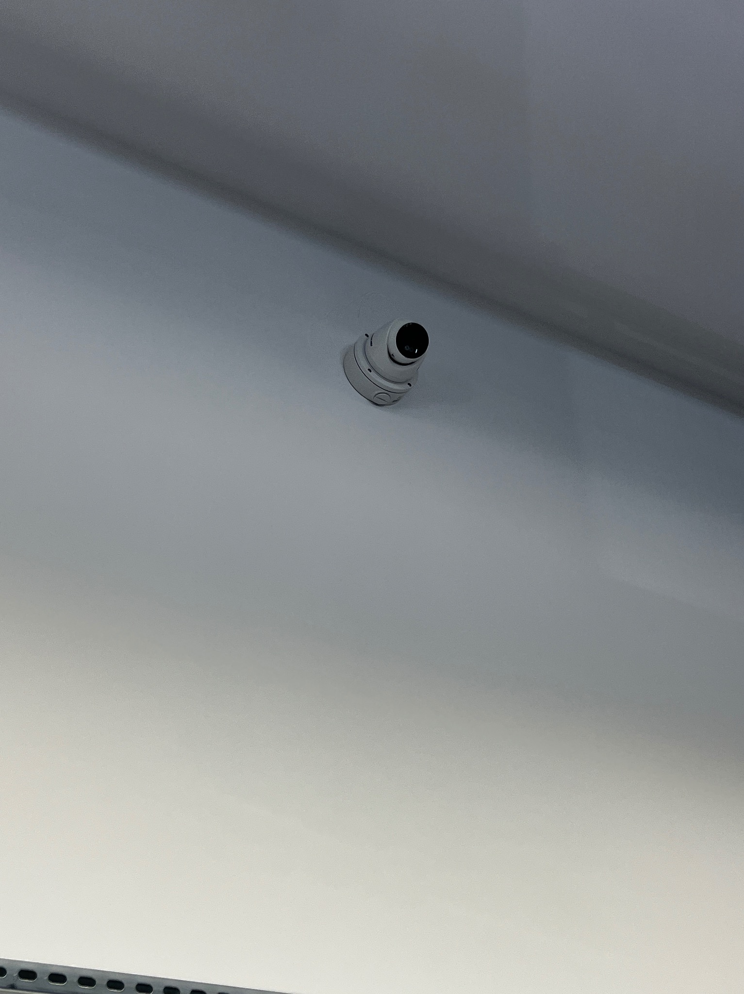 Kamera kopułkowa monitoringu zamontowana na białym suficie, widoczna z dołu pod kątem. Minimalistyczny design kamery, jasne oświetlenie.