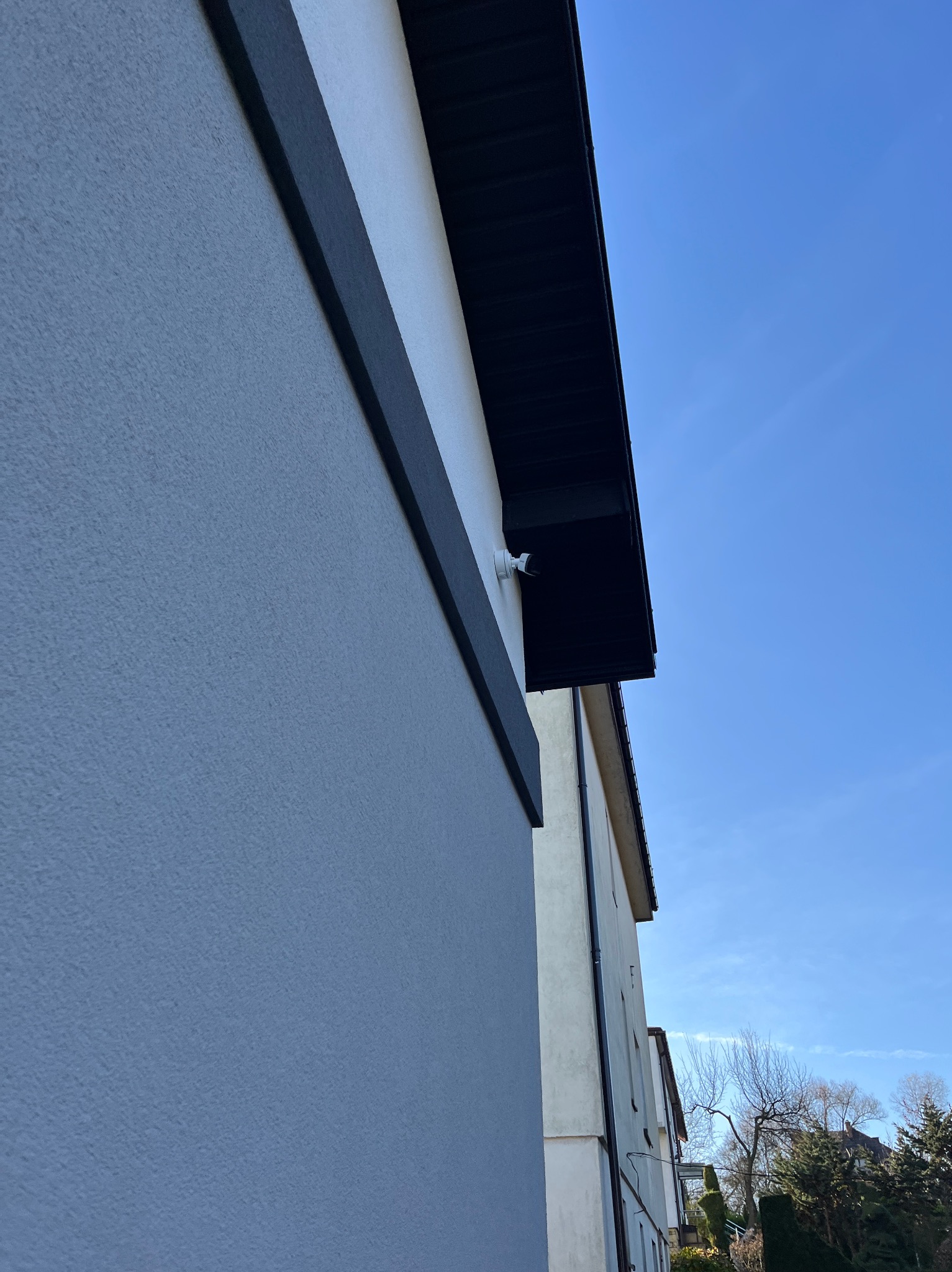 Kamera monitoringu zamontowana na elewacji budynku, widoczna z dołu pod kątem. Biała kamera na tle błękitnego nieba i fragmentu ściany budynku.