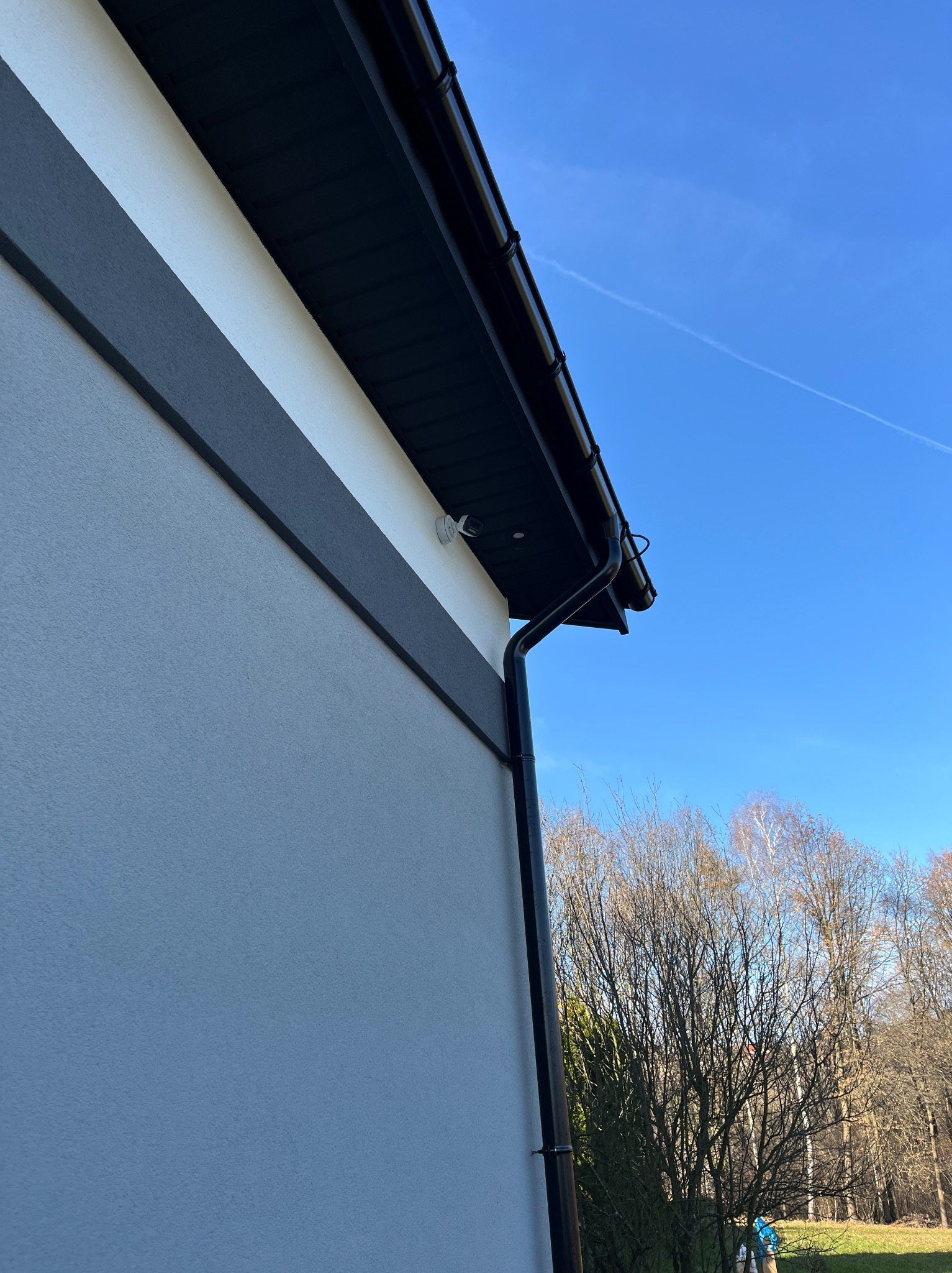 Kamera monitoringu zamontowana na elewacji domu z czarną rynną, widoczna na tle błękitnego nieba i drzew bez liści. Ujęcie z dołu, podkreślające nowoczesny design.