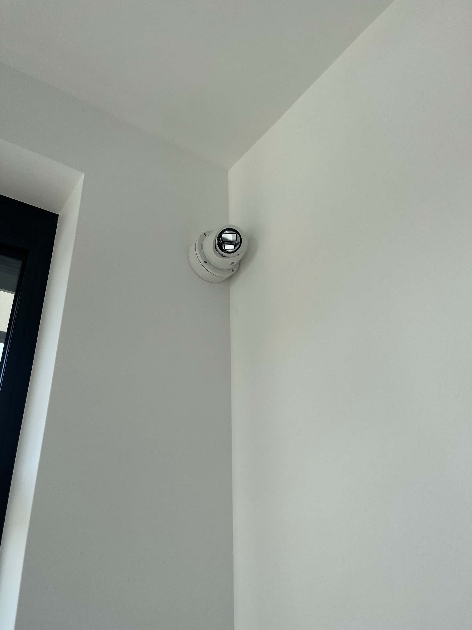 Kamera kopułkowa monitoringu, zamontowana w rogu pomieszczenia z białymi ścianami, obok ramy okiennej. Widok z dołu, oświetlenie dzienne.