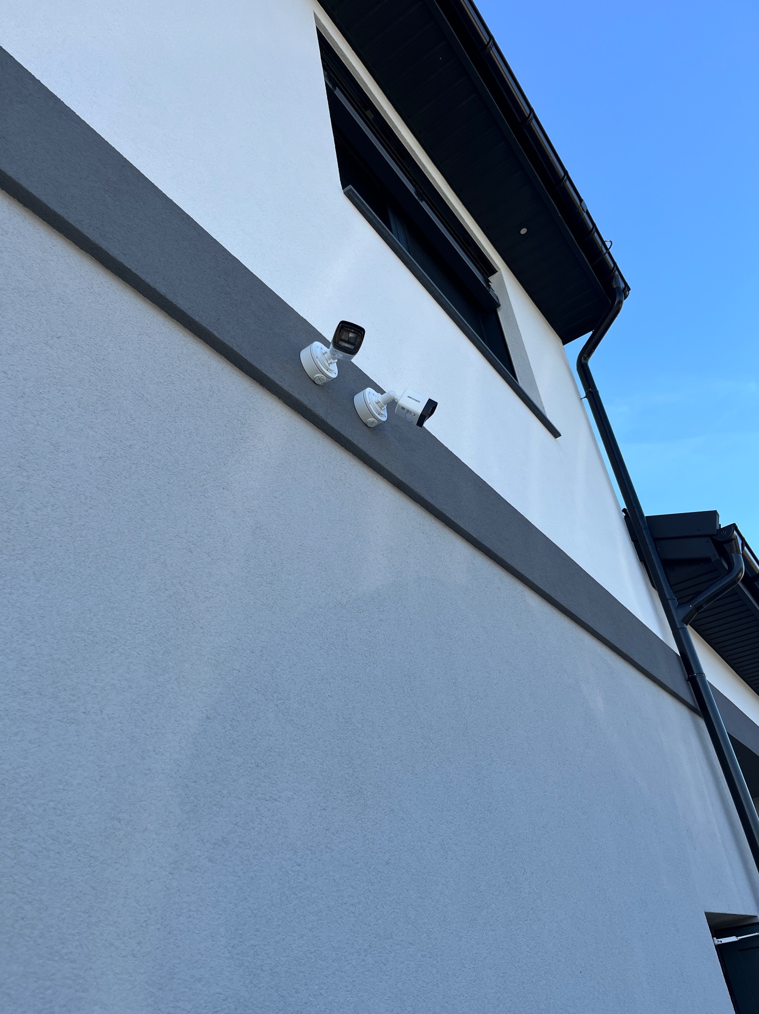 Białe kamery monitoringu Hikvision zamontowane na elewacji domu z szarą listwą dekoracyjną i czarną rynną na tle błękitnego nieba. Ujęcie z dołu.