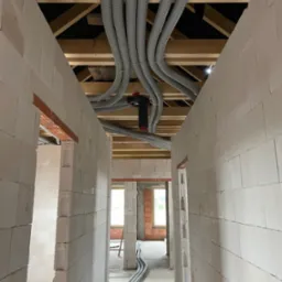 Wnętrze budynku w trakcie budowy z widocznymi szarymi, karbowanymi rurami instalacyjnymi rozprowadzonymi pod drewnianym stropem, prowadzącymi wzdłuż korytarza pomiędzy ścianami z białych bloczków.