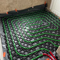 Instal Koncept Artur Biegun - Wnętrze w trakcie montażu ogrzewania podłogowego: zielone rurki ułożone spiralnie na czarnej folii z systemem mocowań, czerwone izolowane końcówki rur, ściany w stanie surowym.