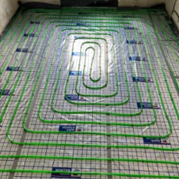 Zielone rury ogrzewania podłogowego ułożone na folii z nadrukiem siatki i logo 'MONTER quality' w surowym wnętrzu, przed wylewką.