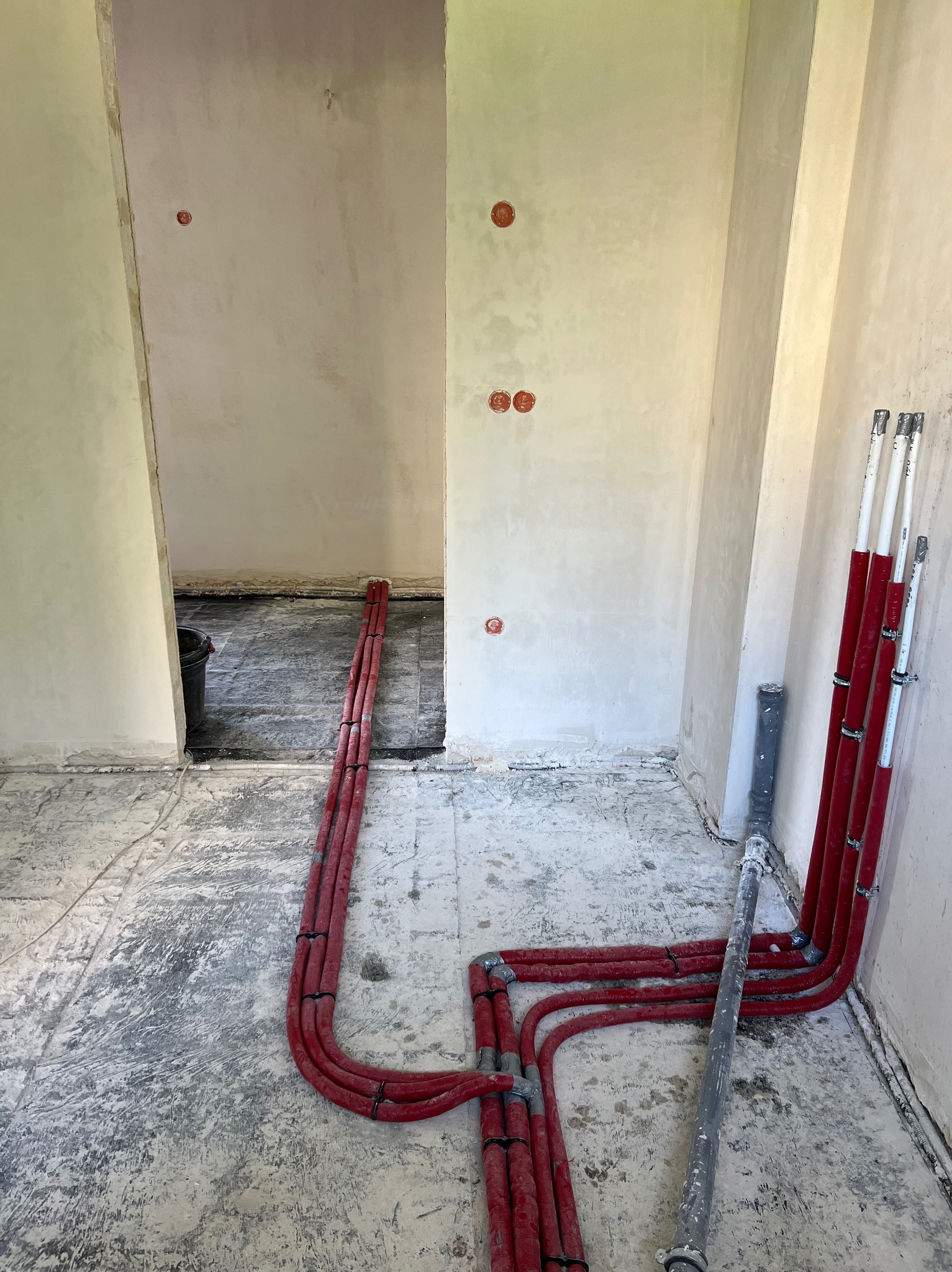 Wnętrze w trakcie remontu z widocznymi czerwonymi rurami instalacji grzewczej układanymi na podłodze, szara rura kanalizacyjna i otwory pod gniazdka w ścianie.