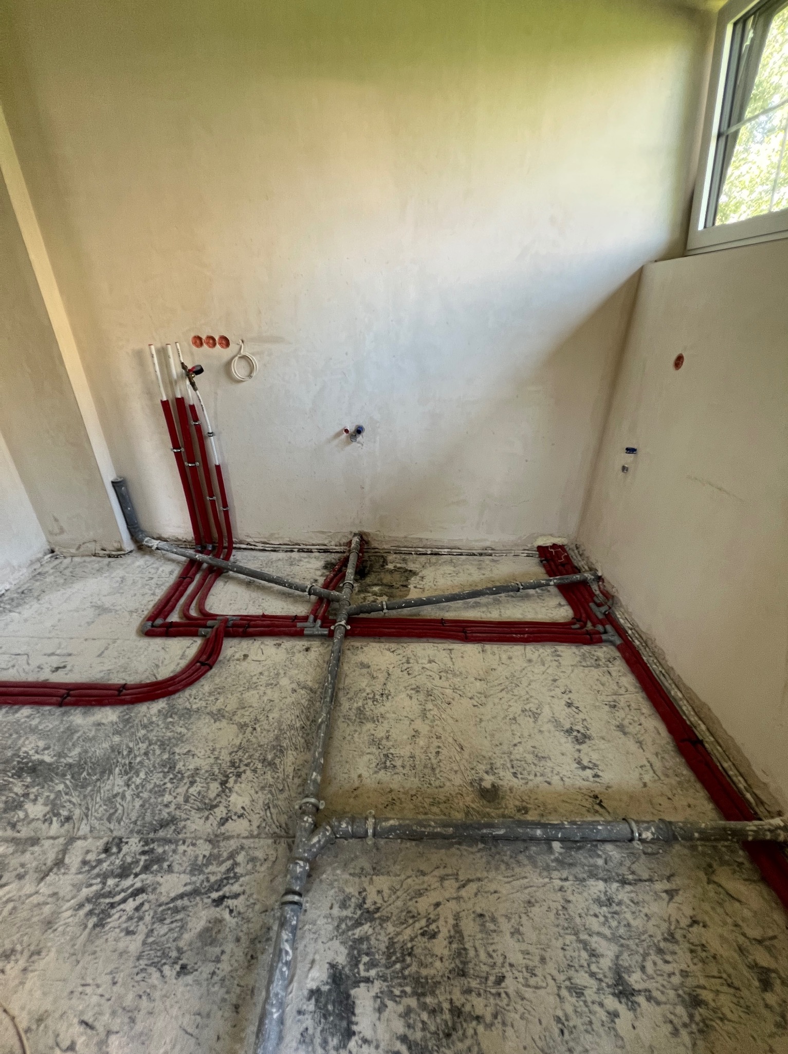 Surowe wnętrze z instalacją wodną i grzewczą w trakcie montażu: czerwone rury PEX prowadzone po podłodze i wzdłuż ściany, szare rury metalowe, widoczne otwory w ścianie pod armaturę i osprzęt.