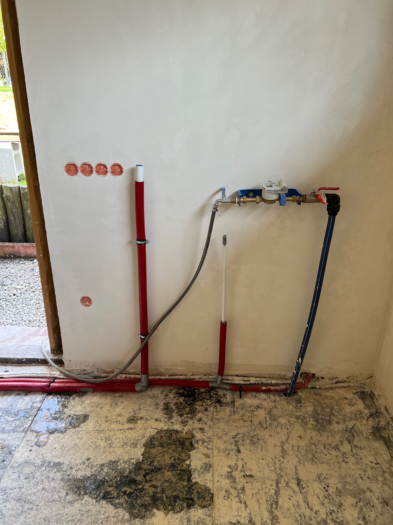 Wnętrze w trakcie prac hydraulicznych: widoczne czerwone i niebieskie rury, licznik wody na ścianie, wywiercone otwory pod instalację elektryczną, surowy stan podłogi i ścian.