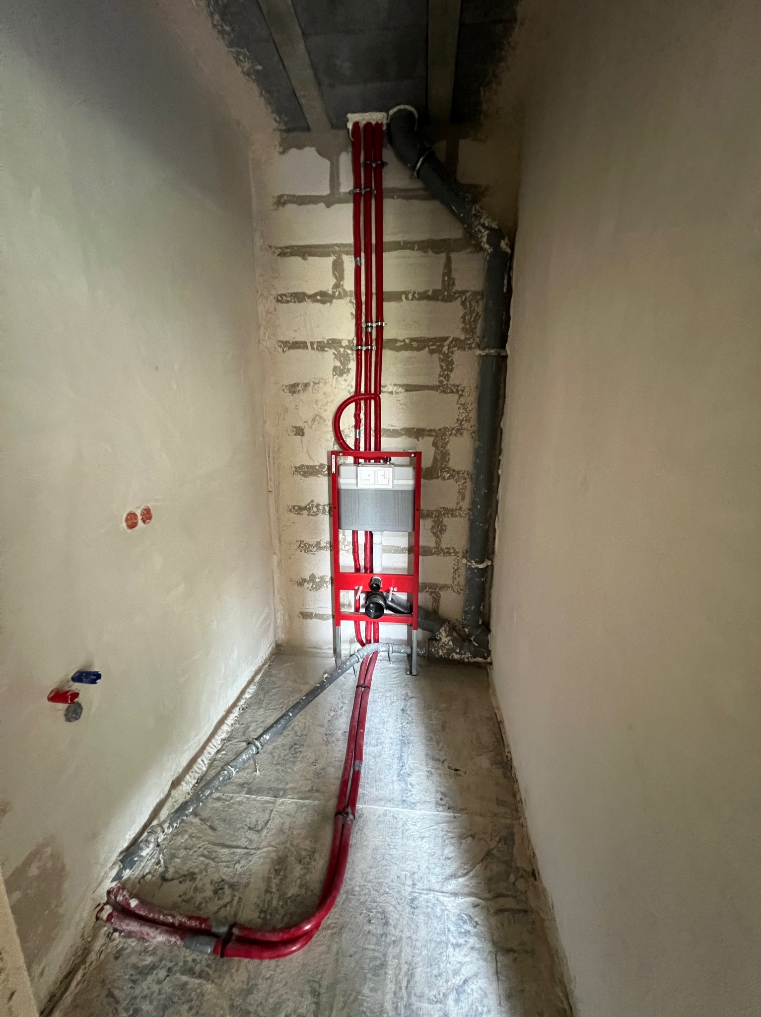 Wąskie pomieszczenie w trakcie remontu z widoczną instalacją podtynkową WC, czerwonymi rurami doprowadzającymi wodę oraz szarą rurą kanalizacyjną.
