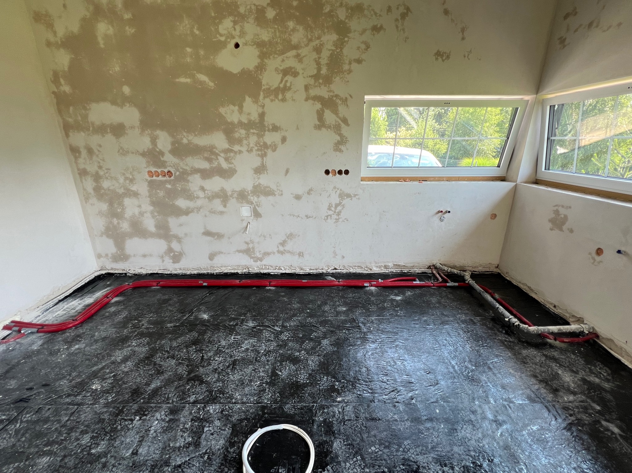 Czerwone rury ogrzewania podłogowego rozłożone na czarnej folii izolacyjnej w surowym pomieszczeniu z oknami dachowymi i ścianami w trakcie wykańczania.