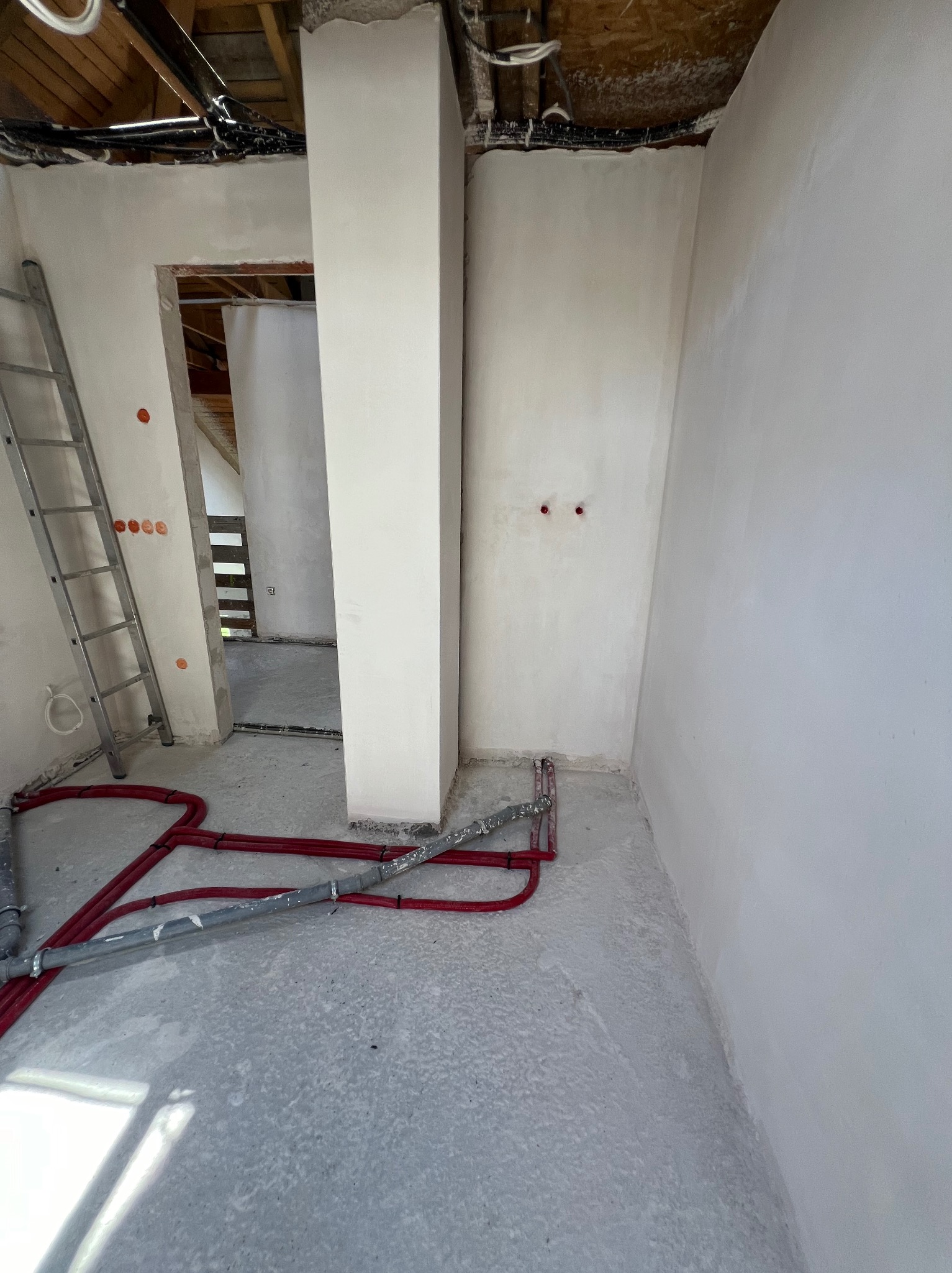 Wnętrze w trakcie remontu z widocznymi czerwonymi rurami instalacji podłogowej, aluminiową drabiną opartą o ścianę i surowym wykończeniem ścian oraz podłogi.