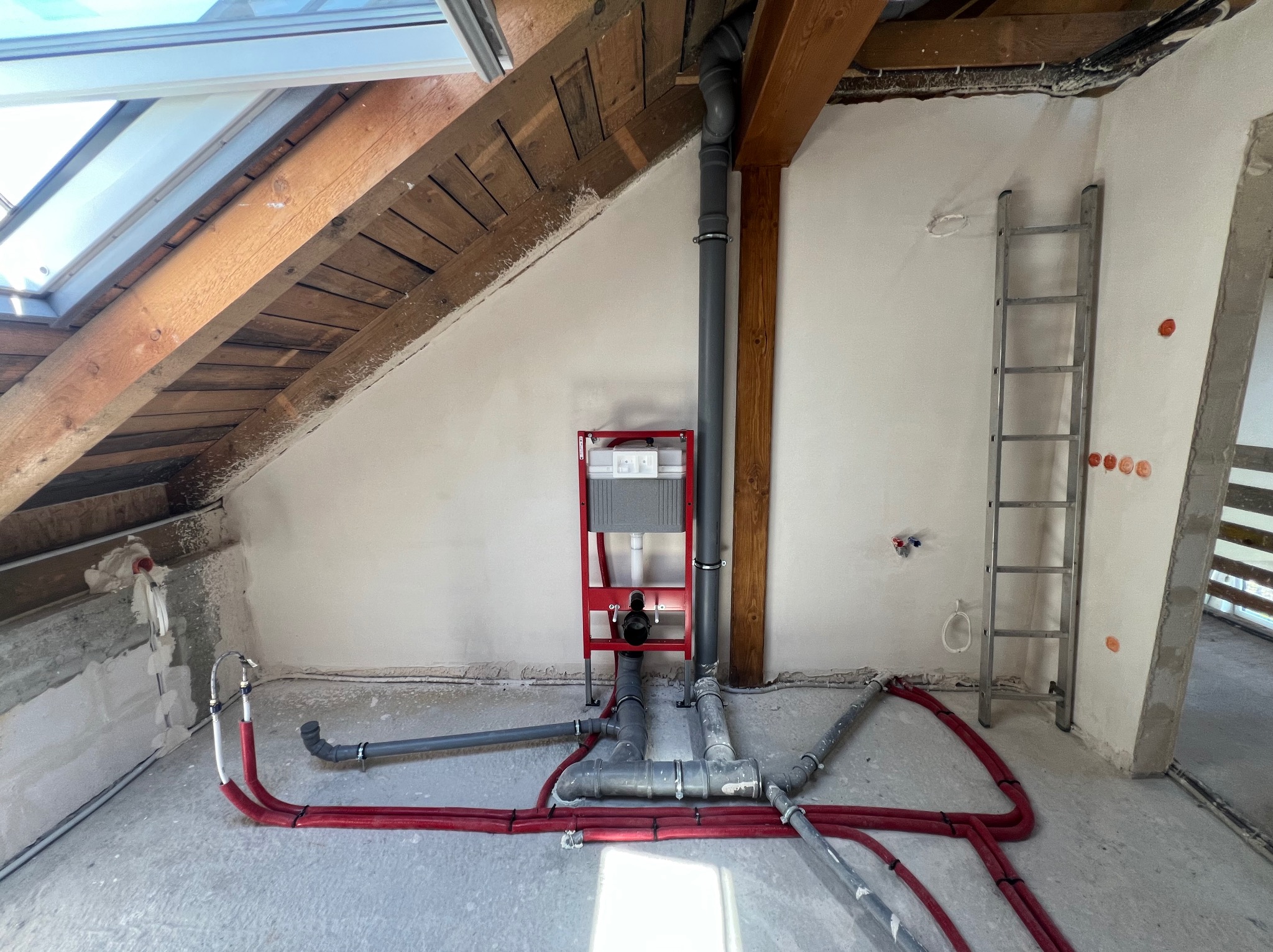 Instalacja w poddaszu: rury grzewcze w czerwonej izolacji, szara rura kanalizacyjna, stelaż podtynkowy WC na czerwonej ramie, drewniana więźba dachowa, drabina oparta o ścianę.