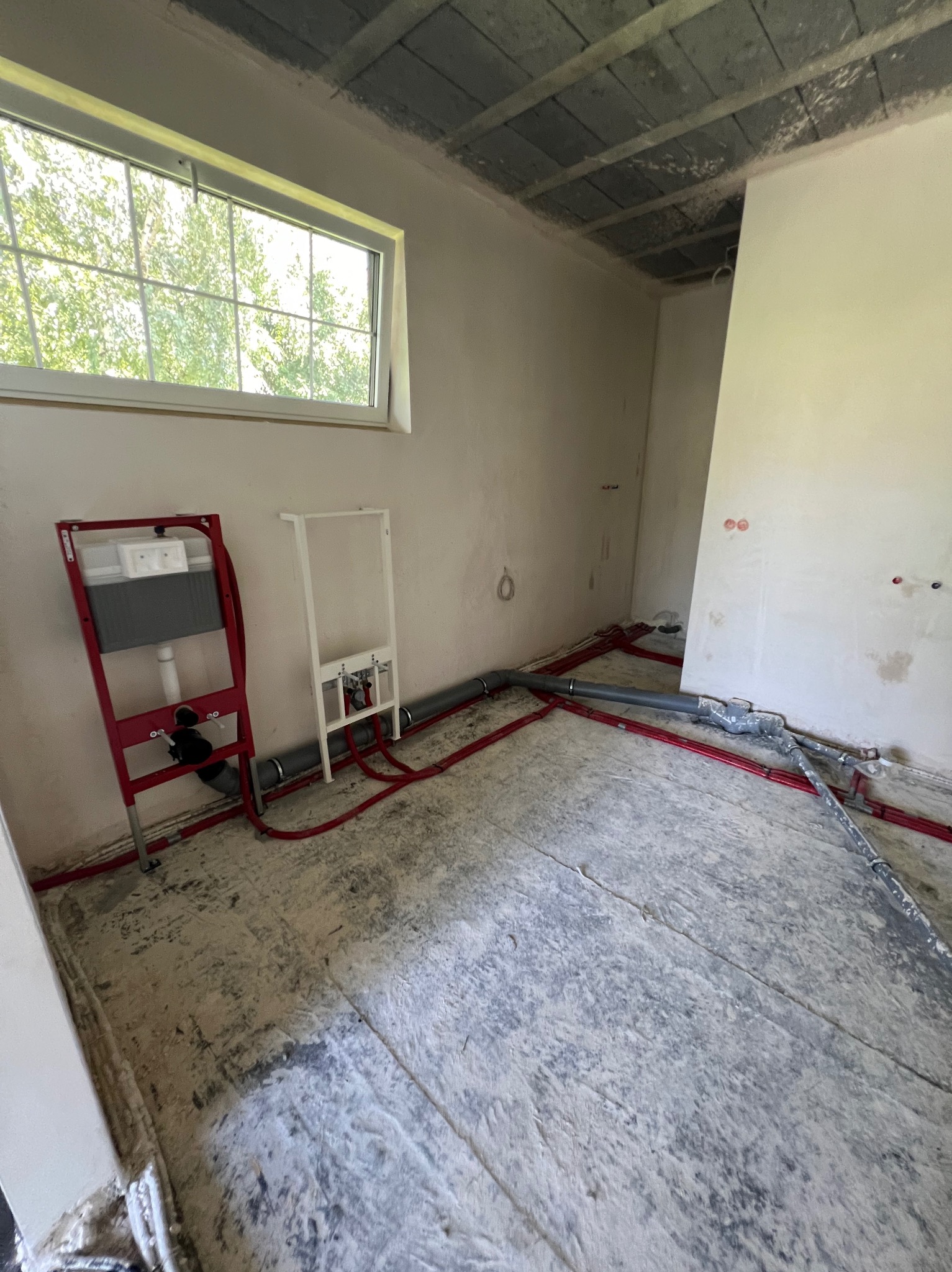 Surowy stan łazienki w trakcie montażu instalacji wodno-kanalizacyjnej: widoczne stelaże podtynkowe WC, rury w kolorze czerwonym i szarym prowadzone po podłodze, przygotowane punkty przyłączeniowe...
