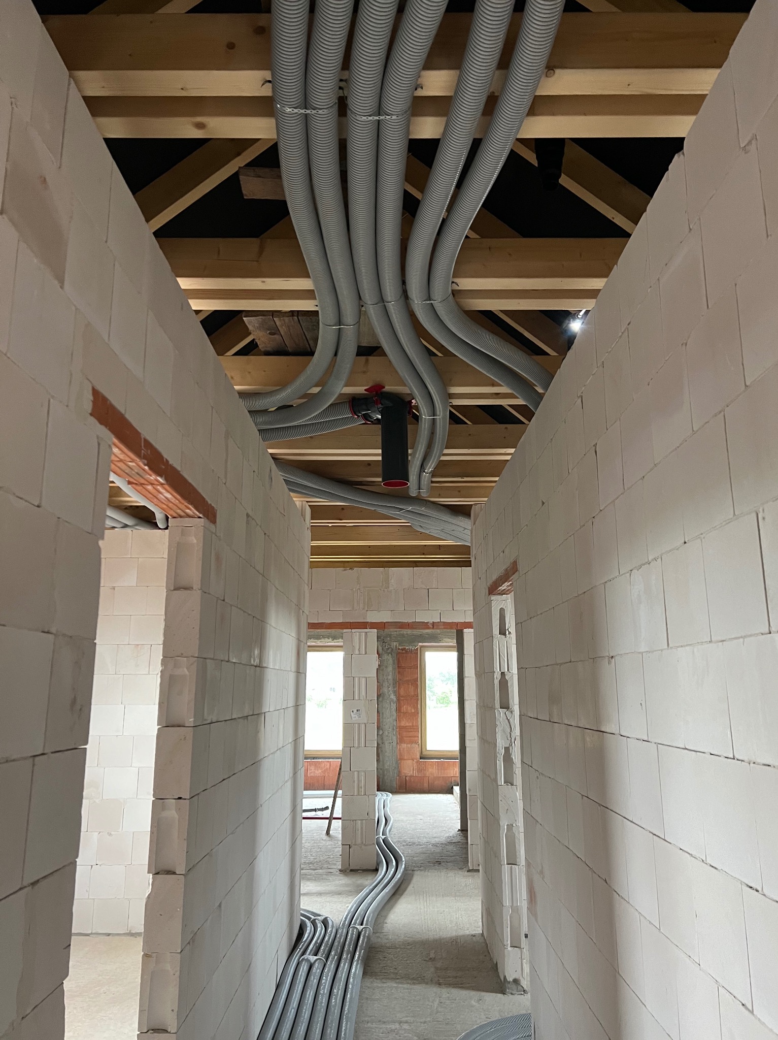 Wnętrze budynku w trakcie budowy z widocznymi szarymi, karbowanymi rurami instalacyjnymi rozprowadzonymi pod drewnianym stropem, prowadzącymi wzdłuż korytarza pomiędzy ścianami z białych bloczków.