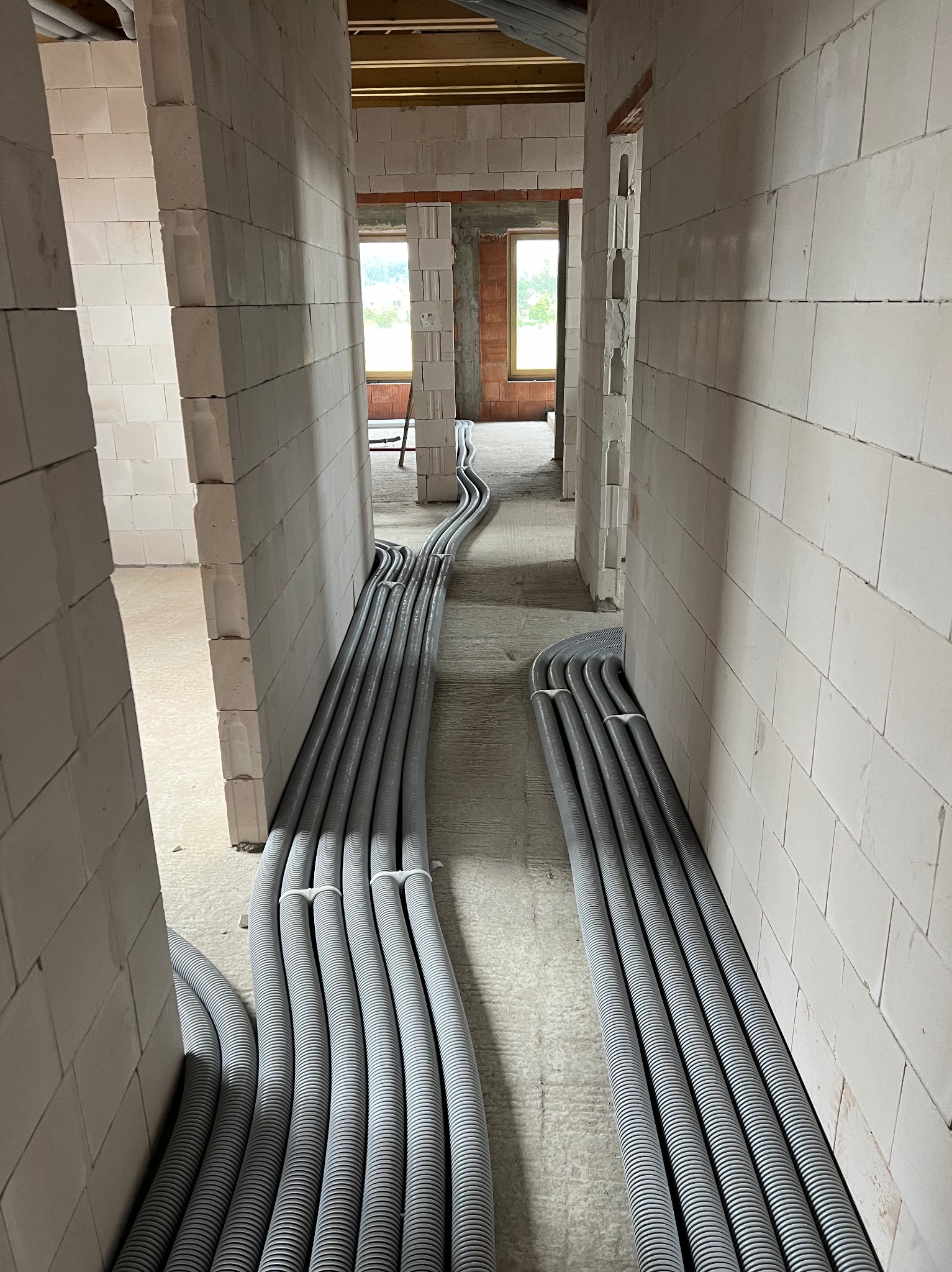 Szereg szarych, karbowanych rur instalacyjnych ułożonych równolegle w surowym wnętrzu budynku z bloczków, widoczne okna w tle.