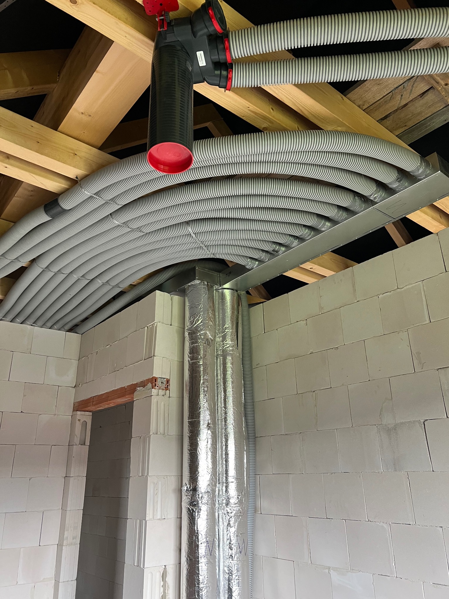Szereg szarych, karbowanych rur wentylacyjnych ułożonych równolegle pod drewnianym stropem, z czarnym rozdzielaczem z czerwonymi elementami, oraz pionowa rura owinięta srebrną folią, widoczne...