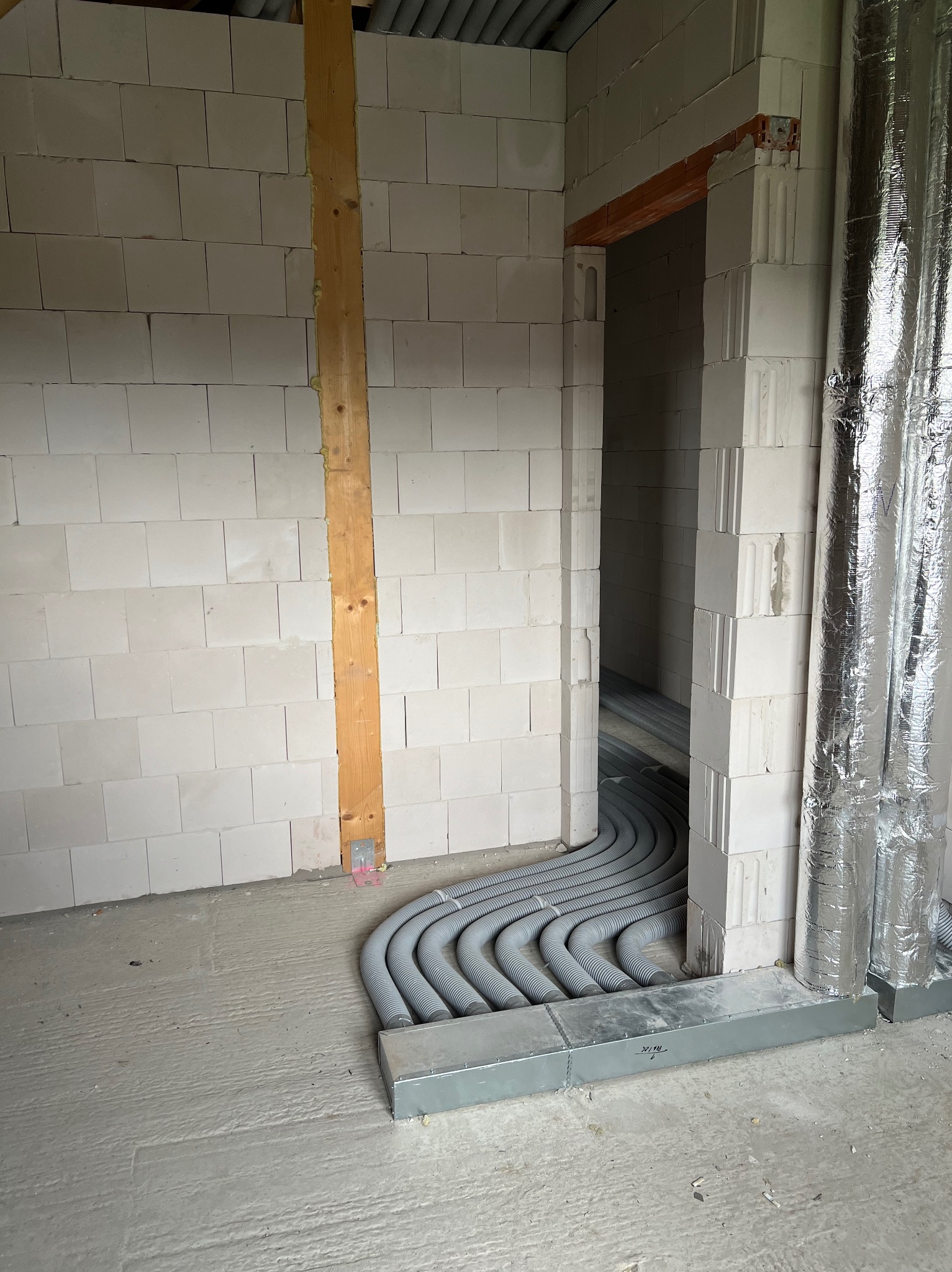 Wnętrze w trakcie budowy z widocznymi rurami instalacji podłogowej ułożonymi w rowkach w posadzce, ściany z białych bloczków i izolowany przewód wentylacyjny.