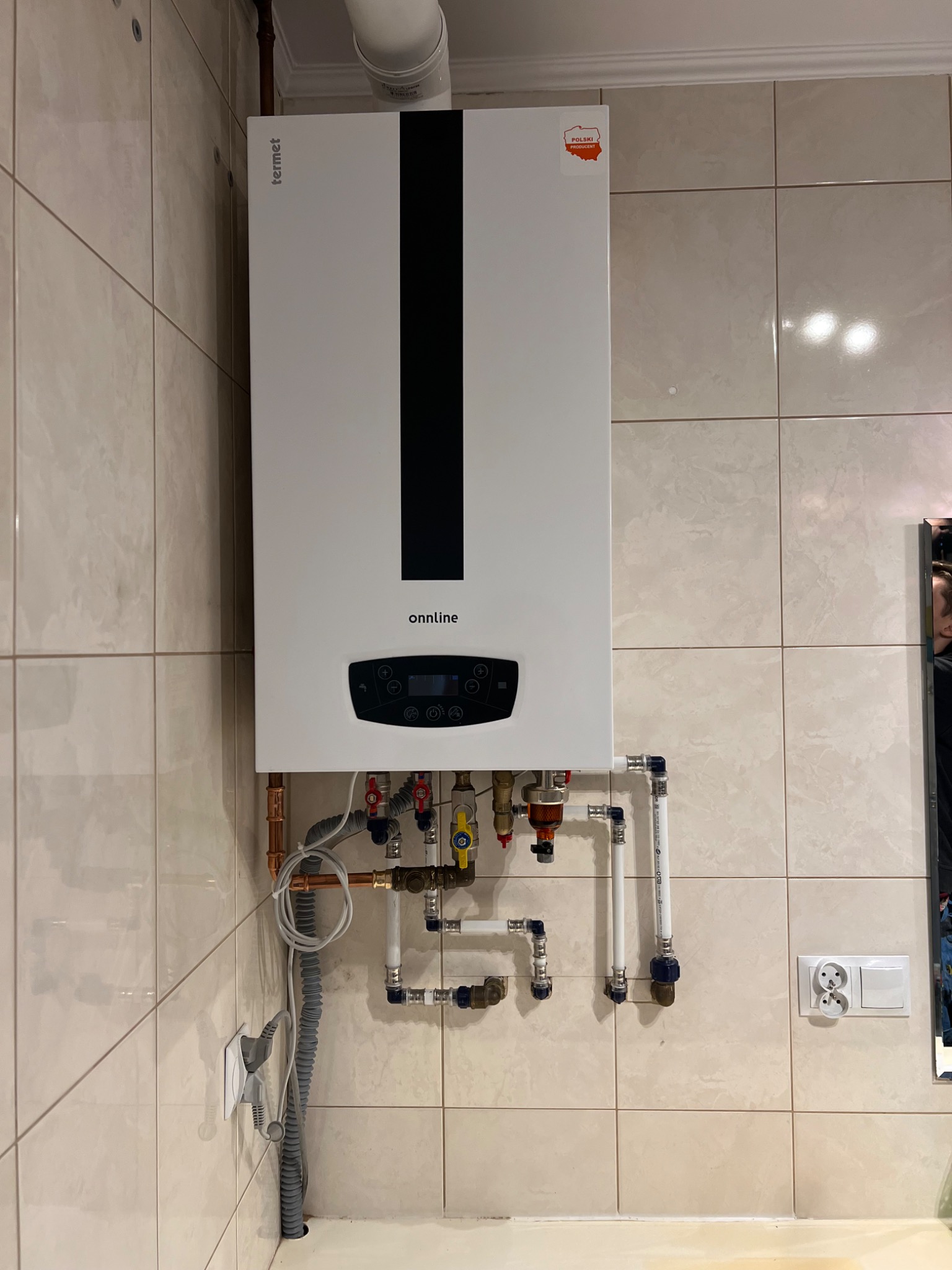 Biały, wiszący piec gazowy marki Termet Onnline na tle beżowych, kwadratowych płytek. Widoczne rury miedziane i plastikowe, zawory, filtr oraz gniazdko elektryczne. Na piecu naklejka 'Polski...