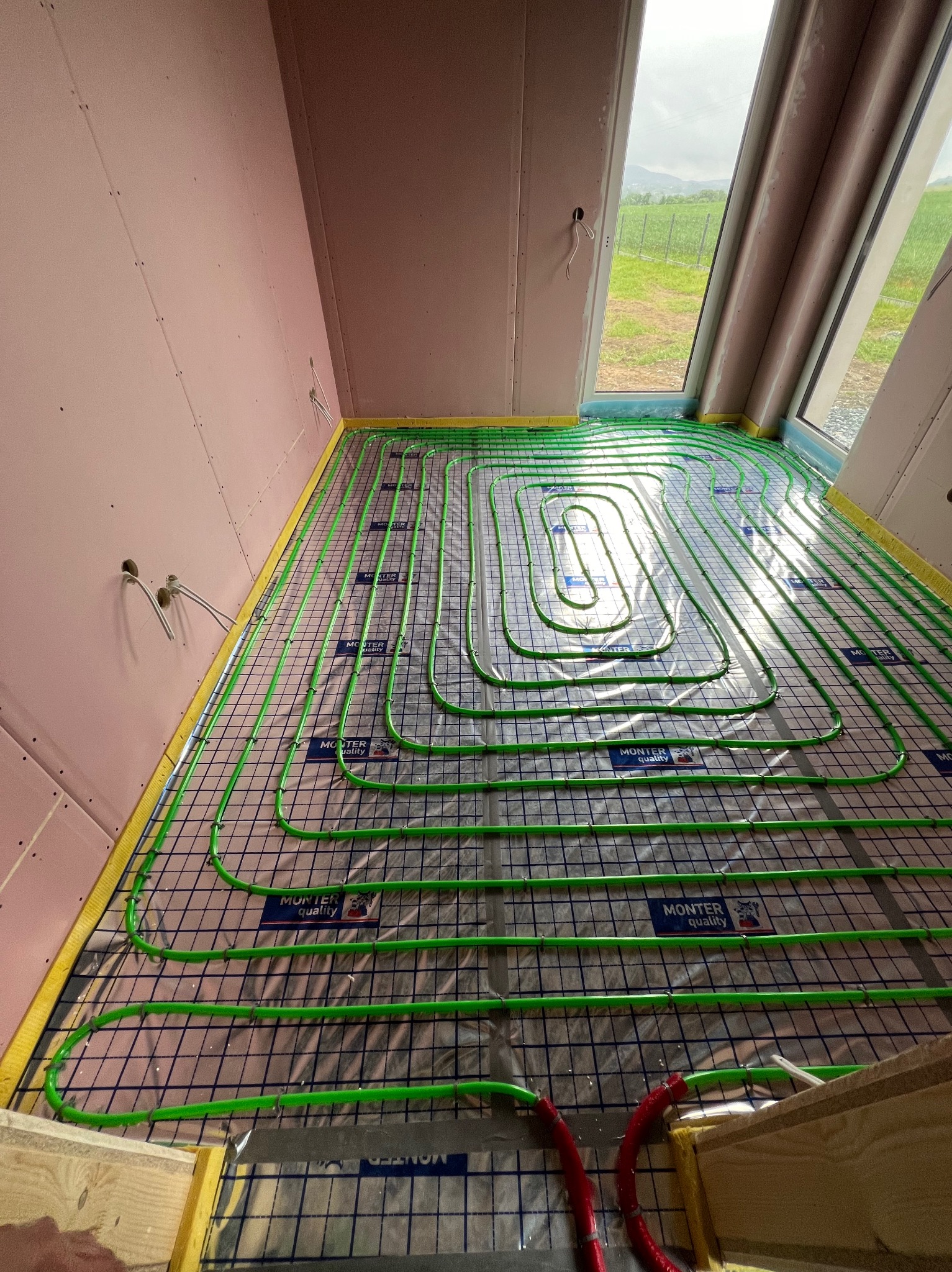 Zielone rury ogrzewania podłogowego ułożone na siatce w surowym wnętrzu z widokiem na zielone wzgórza za oknem; widoczne etykiety 'MONTER quality' na rurach.