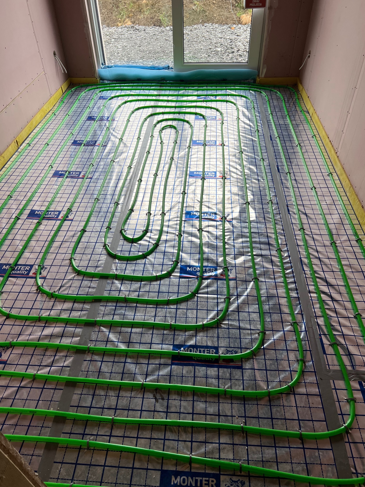 Zielone rurki ogrzewania podłogowego ułożone na folii z siatką, przymocowane plastikowymi klipsami w surowym stanie, widok z góry na przygotowaną instalację przed wylaniem wylewki.