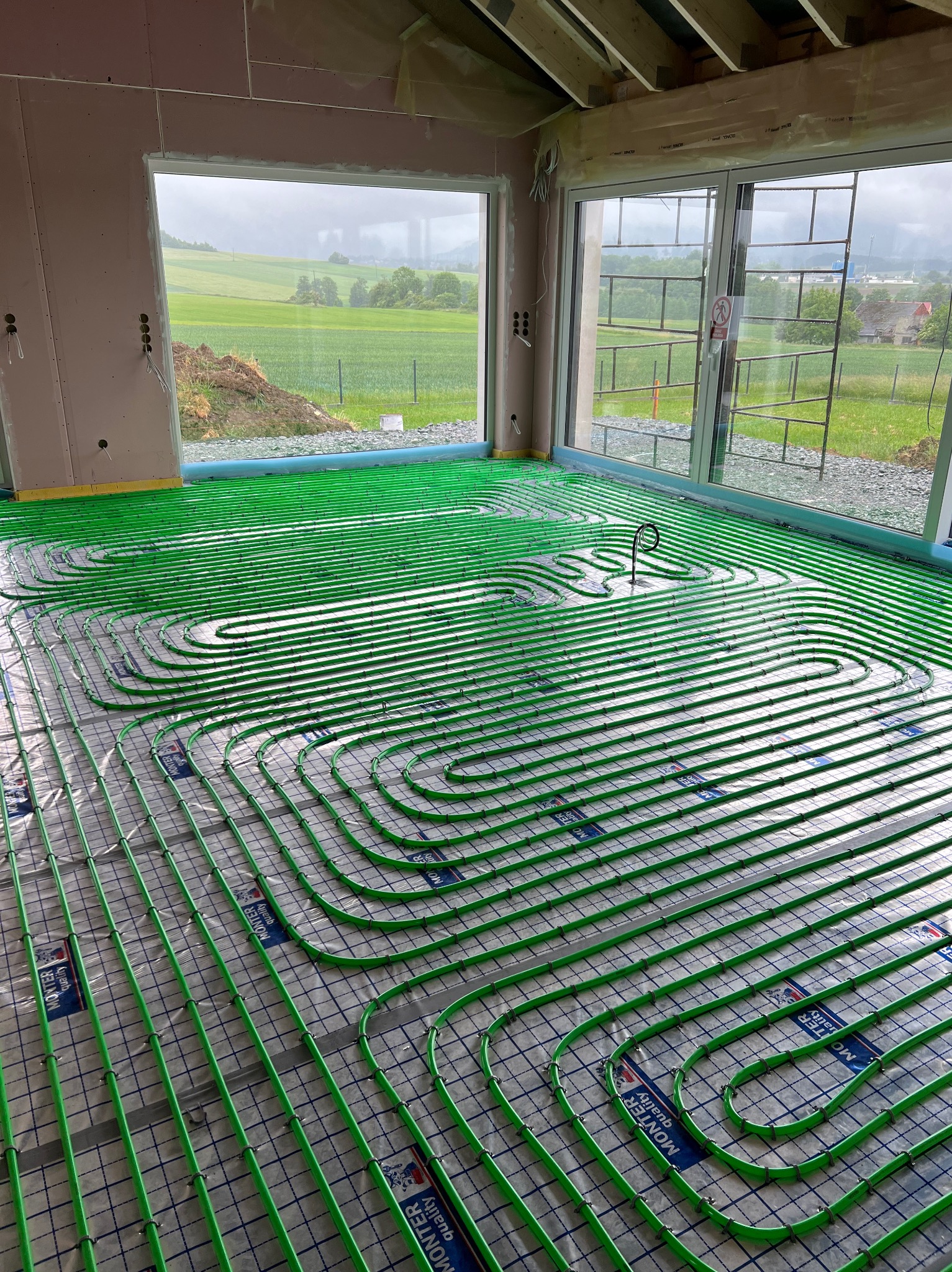 Zielone rury ogrzewania podłogowego ułożone na folii z siatką w surowym wnętrzu domu z widokiem na zielony krajobraz za oknem.