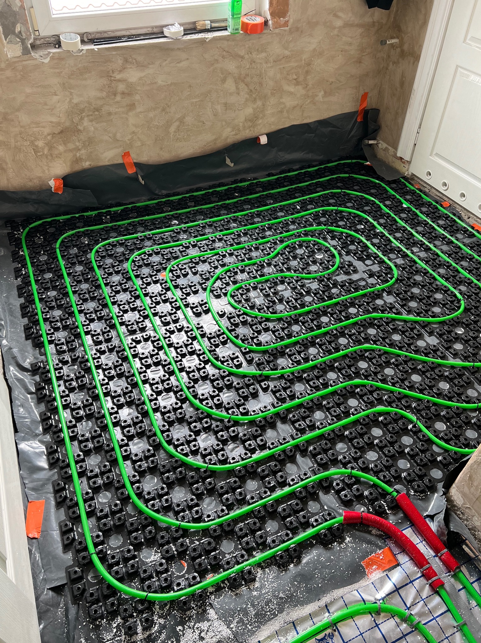 Wnętrze w trakcie montażu ogrzewania podłogowego: zielone rurki ułożone spiralnie na czarnej folii z systemem mocowań, czerwone izolowane końcówki rur, ściany w stanie surowym.