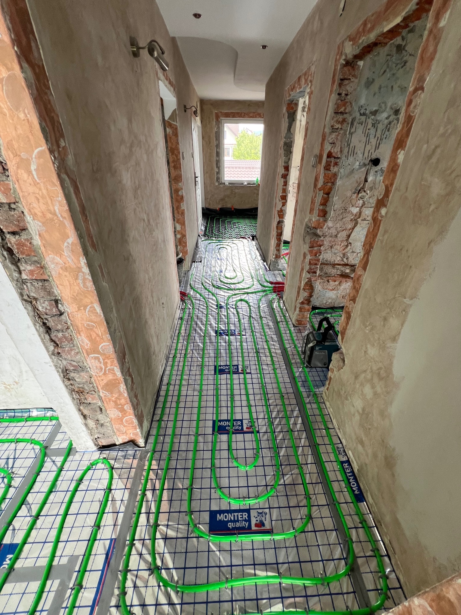 Wnętrze w trakcie remontu z widoczną instalacją ogrzewania podłogowego: zielone rurki ułożone na folii z nadrukiem kratki, surowe ściany z odsłoniętą cegłą, w tle okno z widokiem na zieleń.