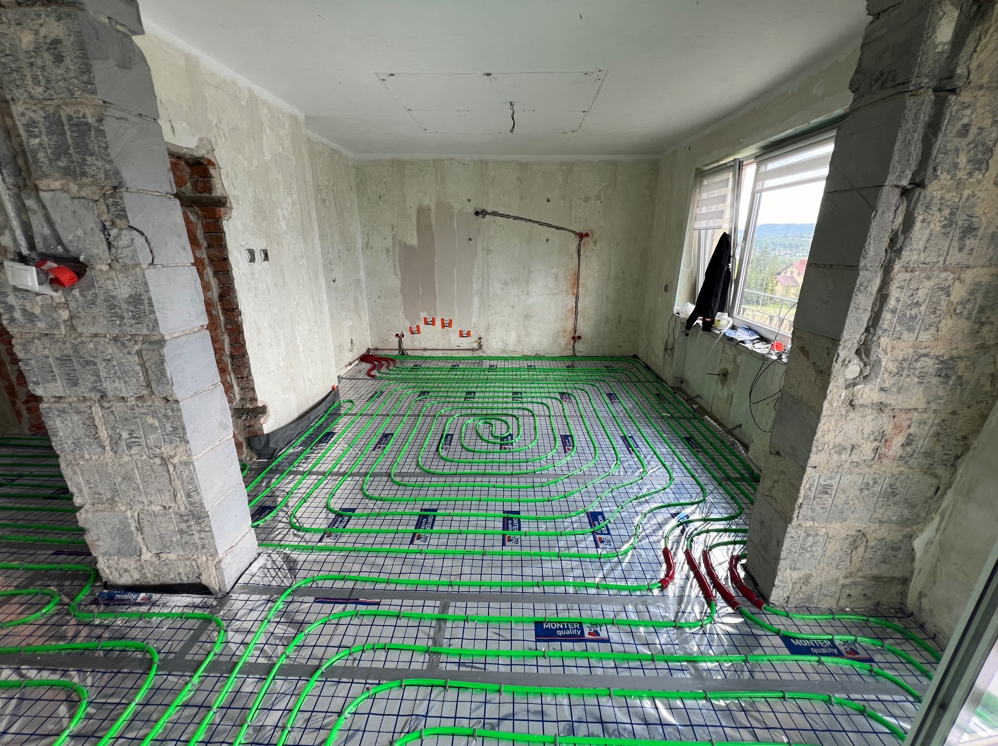 Wnętrze w trakcie remontu z widoczną instalacją ogrzewania podłogowego, zielone rurki ułożone na srebrnej folii z niebieską siatką, surowe ściany i filary, widok na zewnątrz przez okno.