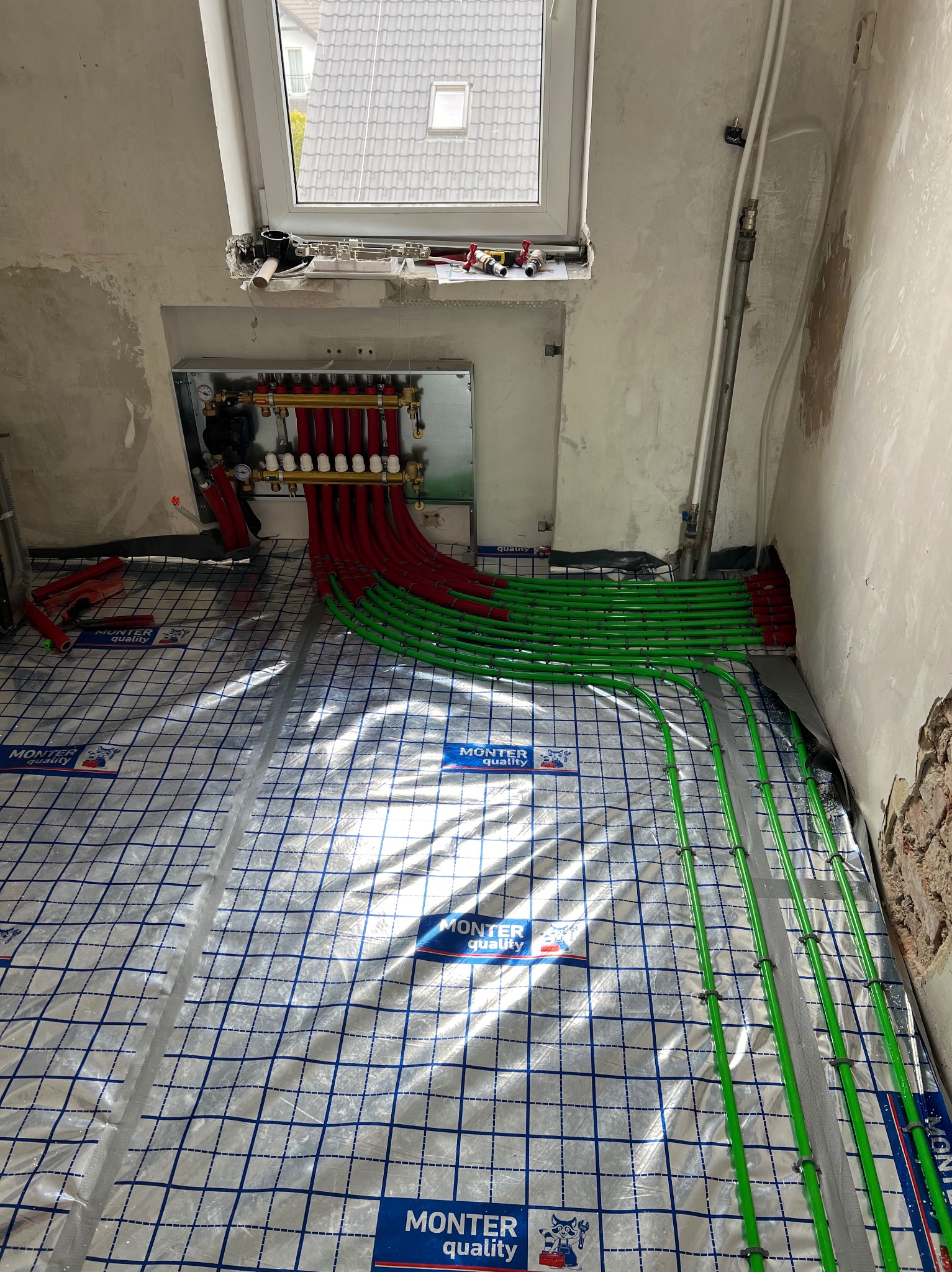 Instalacja ogrzewania podłogowego w surowym wnętrzu, widoczne czerwone i zielone rury rozprowadzone na folii z nadrukiem, kolektor w szarej obudowie, okno dachowe w tle.