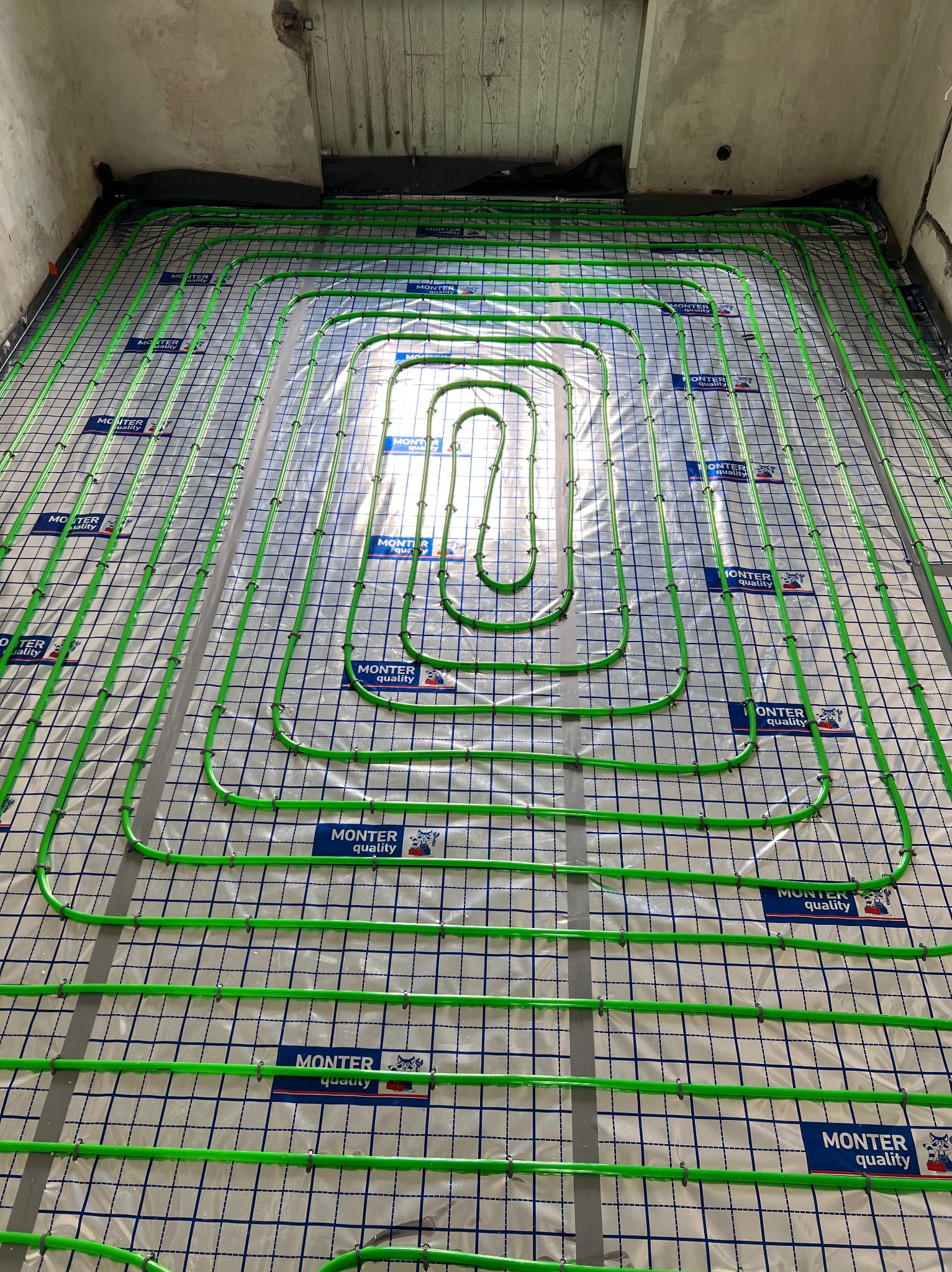 Zielone rury ogrzewania podłogowego ułożone na folii z nadrukiem siatki i logo 'MONTER quality' w surowym wnętrzu, przed wylewką.
