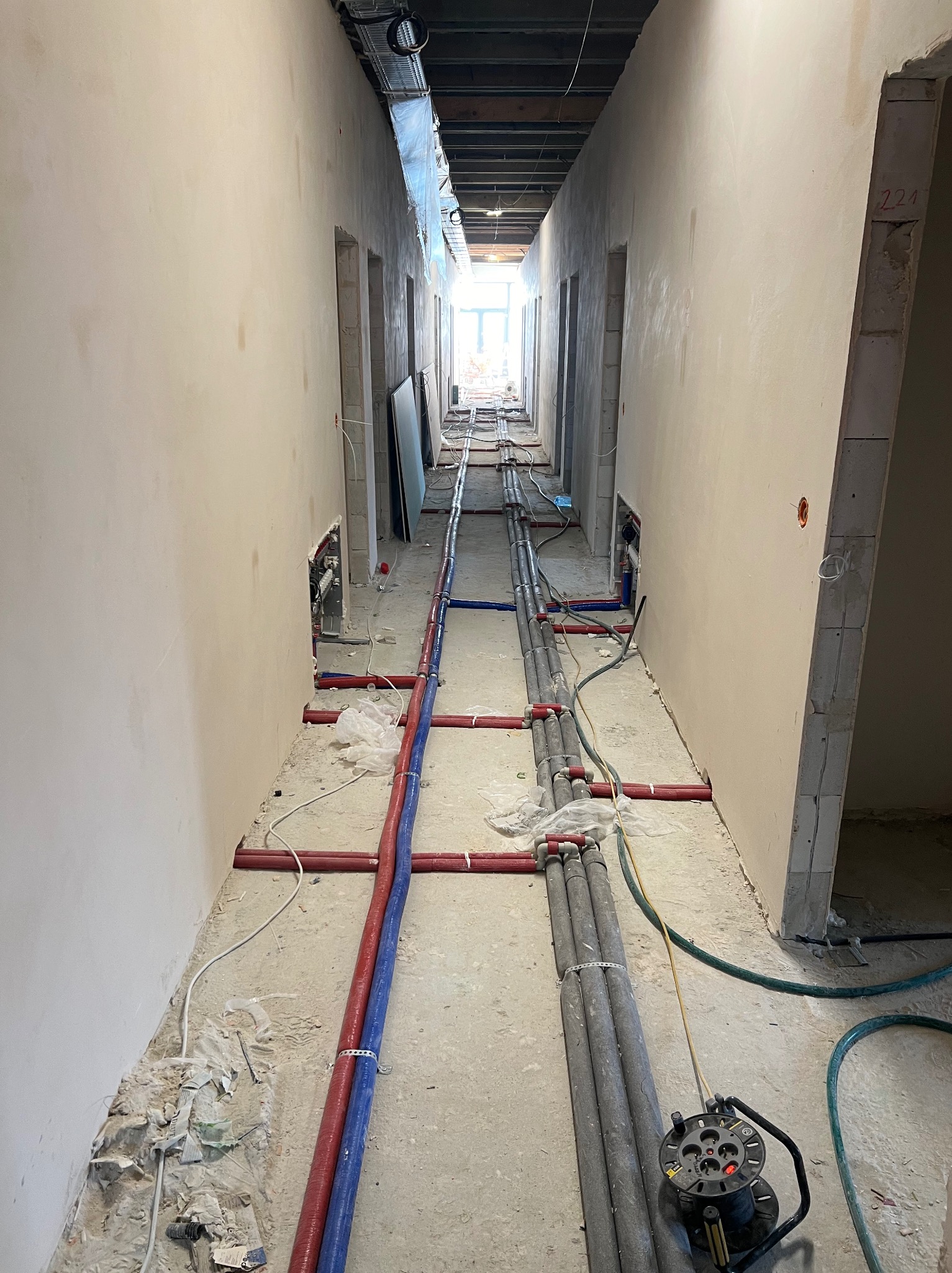 Korytarz w trakcie remontu z widocznymi rurami instalacji hydraulicznej i grzewczej ułożonymi na podłodze, gniazdo elektryczne na zwijaku i surowe ściany w tle.