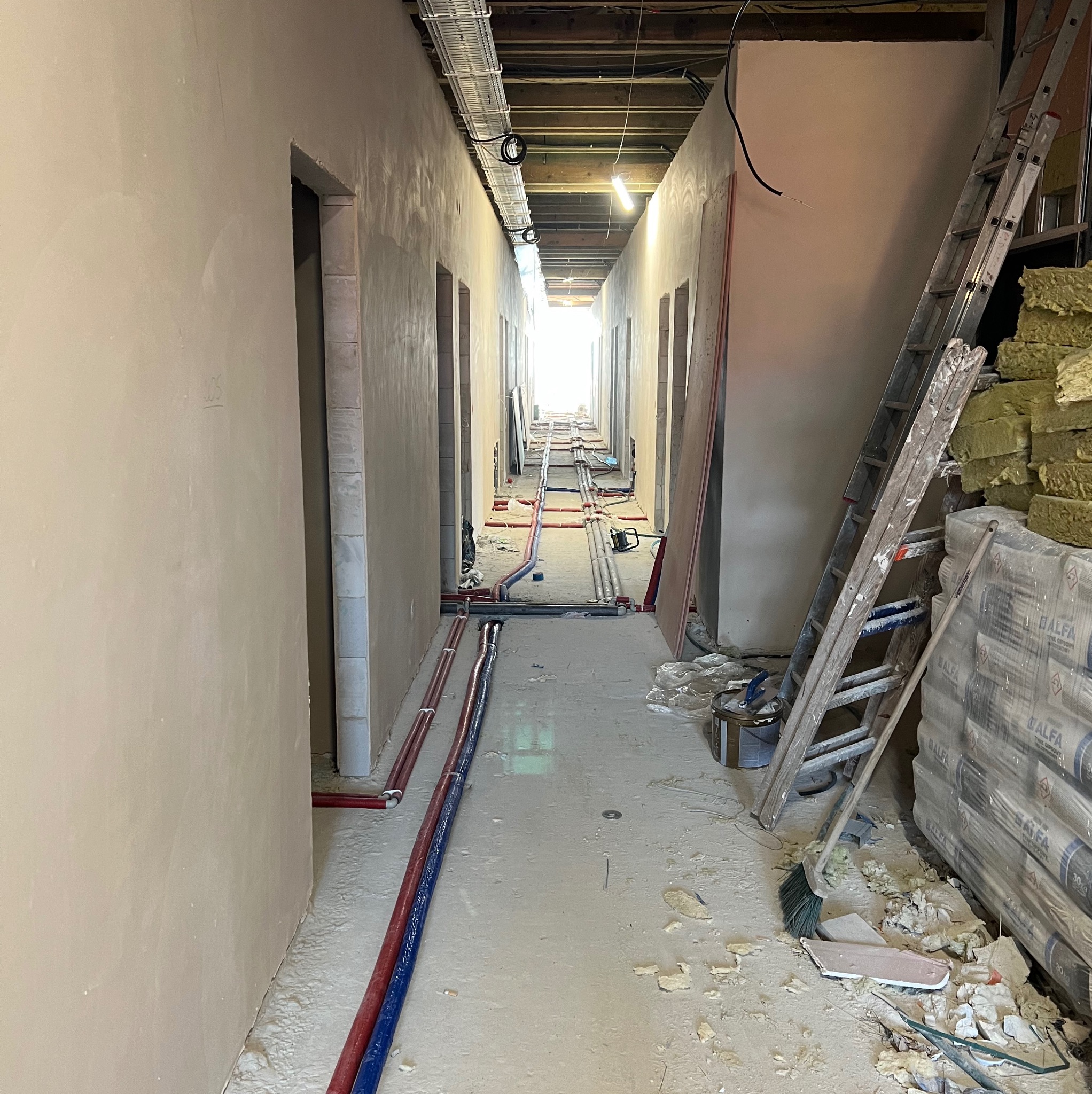 Korytarz w trakcie kompleksowego wykonania instalacji hydraulicznych i grzewczych: widoczne rury w kolorze czerwonym i niebieskim, drabina oparta o ścianę, worki z materiałem izolacyjnym i surowy...