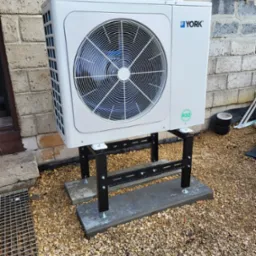 Biała pompa ciepła marki York zamontowana na czarnym, metalowym stelażu na zewnątrz budynku z cegły, otoczona żwirem.
