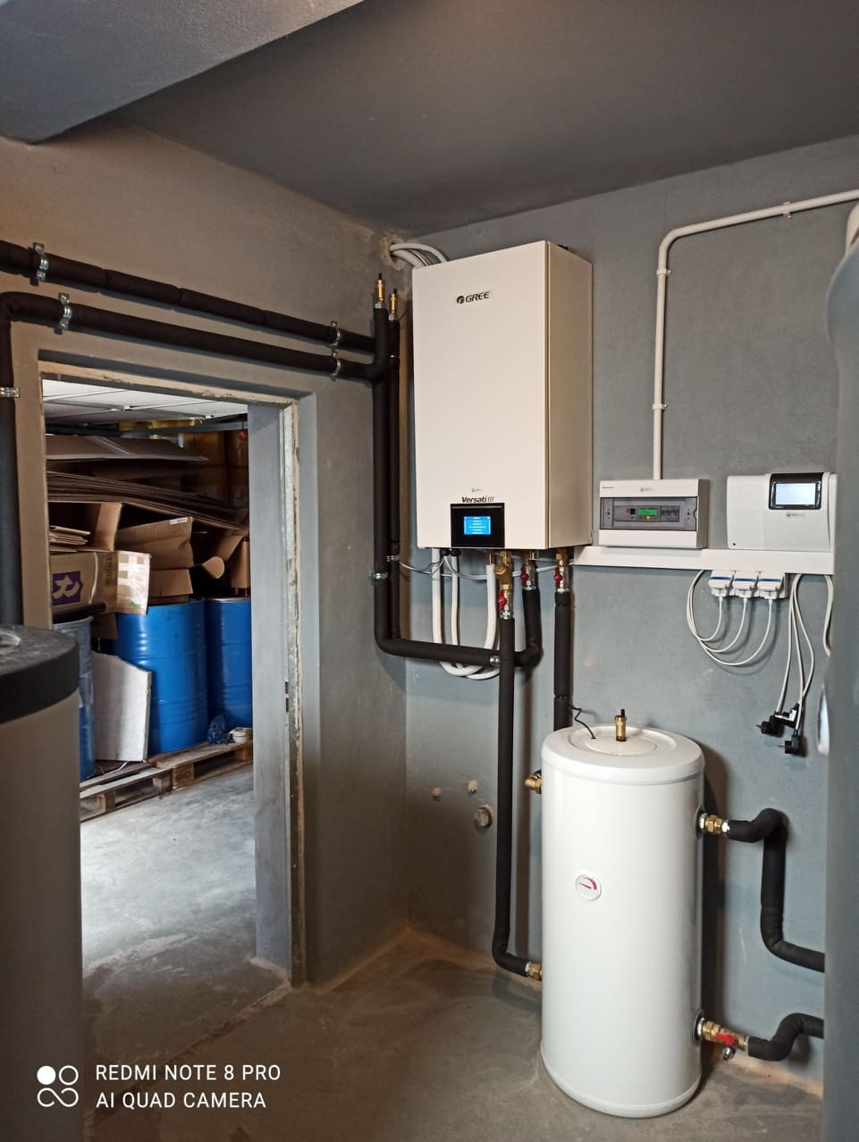 Nowoczesna instalacja z białą jednostką grzewczą Gree Versati III z czarnymi, ocieplonymi rurami, białym zbiornikiem i sterownikiem na szarej ścianie w surowym pomieszczeniu z betonową podłogą...