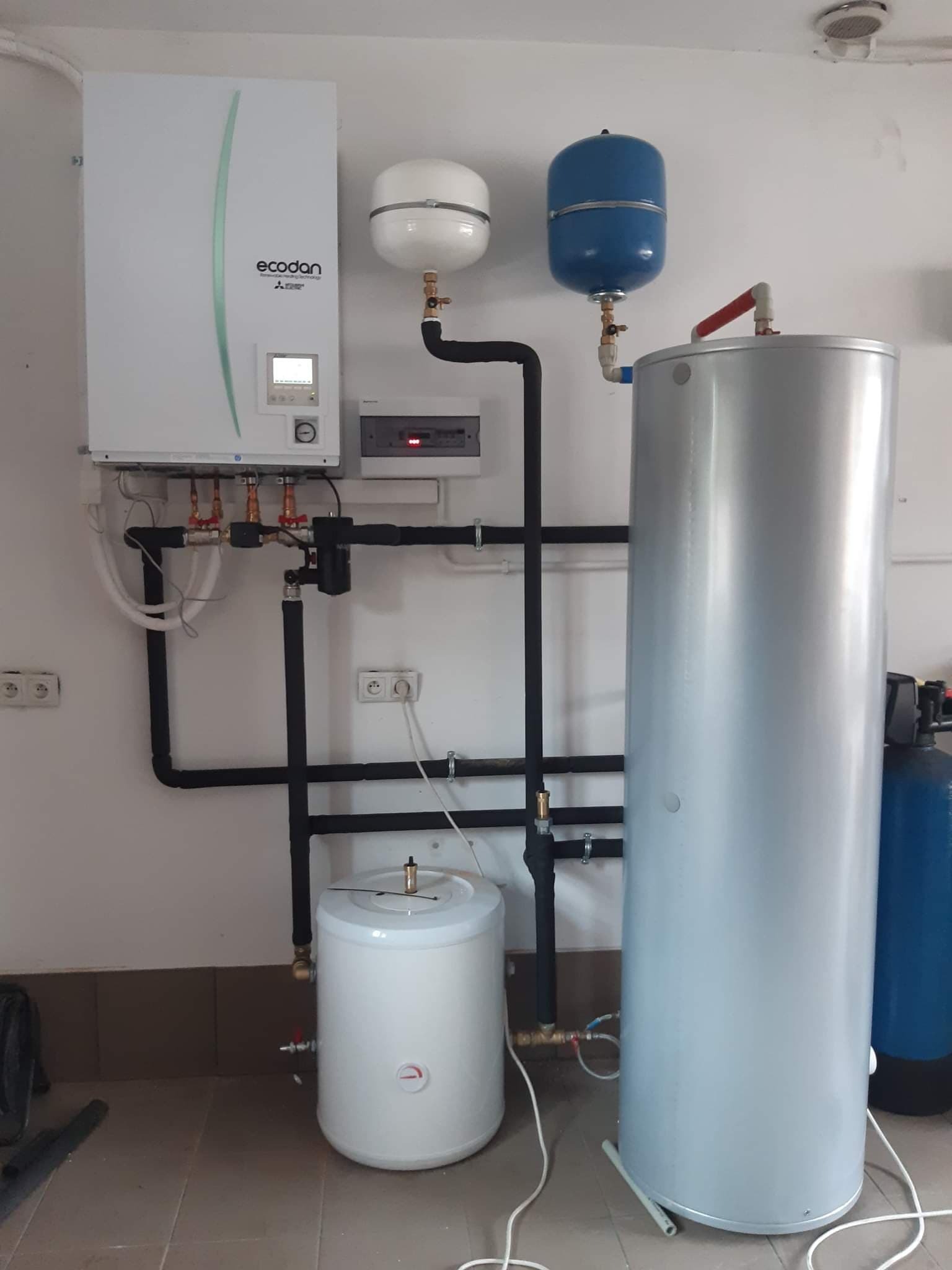 Instalacja kotłowni z białą jednostką Ecodan, zbiornikiem akumulacyjnym i naczyniami wzbiorczymi w pomieszczeniu z brązowymi płytkami na dole ściany.