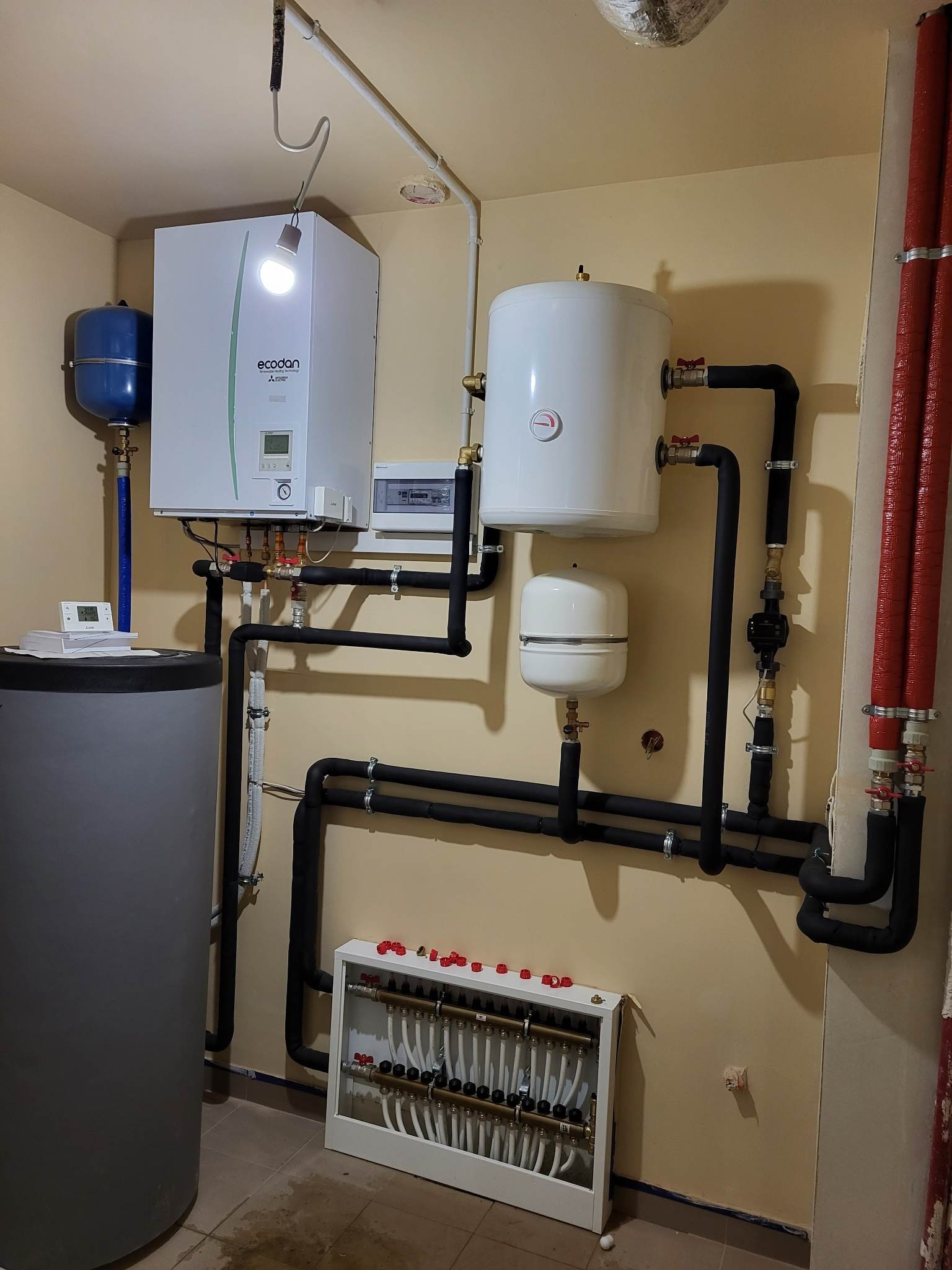 Instalacja grzewcza w kotłowni: zbiornik buforowy, pompa ciepła Ecodan z oświetleniem, dwa zbiorniki wyrównawcze, rozdzielacz ogrzewania podłogowego z licznymi zaworami, rury w izolacji termicznej.