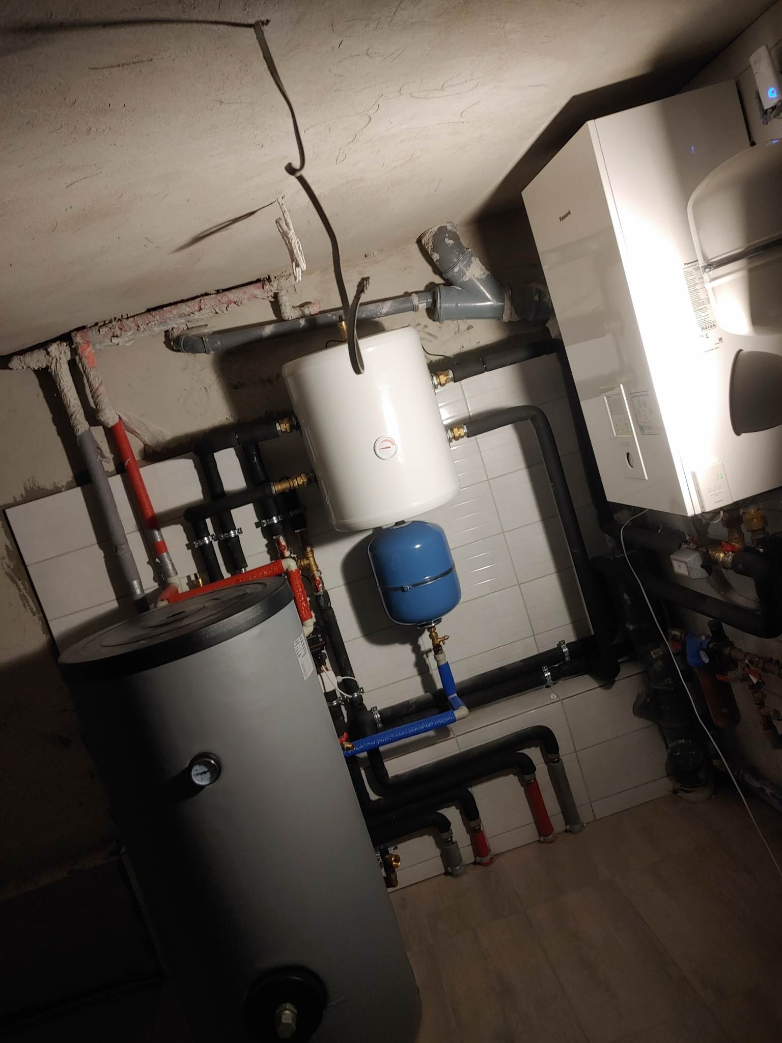 Instalacja z zasobnikiem ciepłej wody, zbiornikiem wyrównawczym i rurami w pomieszczeniu z surowym betonowym sufitem i częściowo wykończonymi ścianami, widoczne białe płytki.