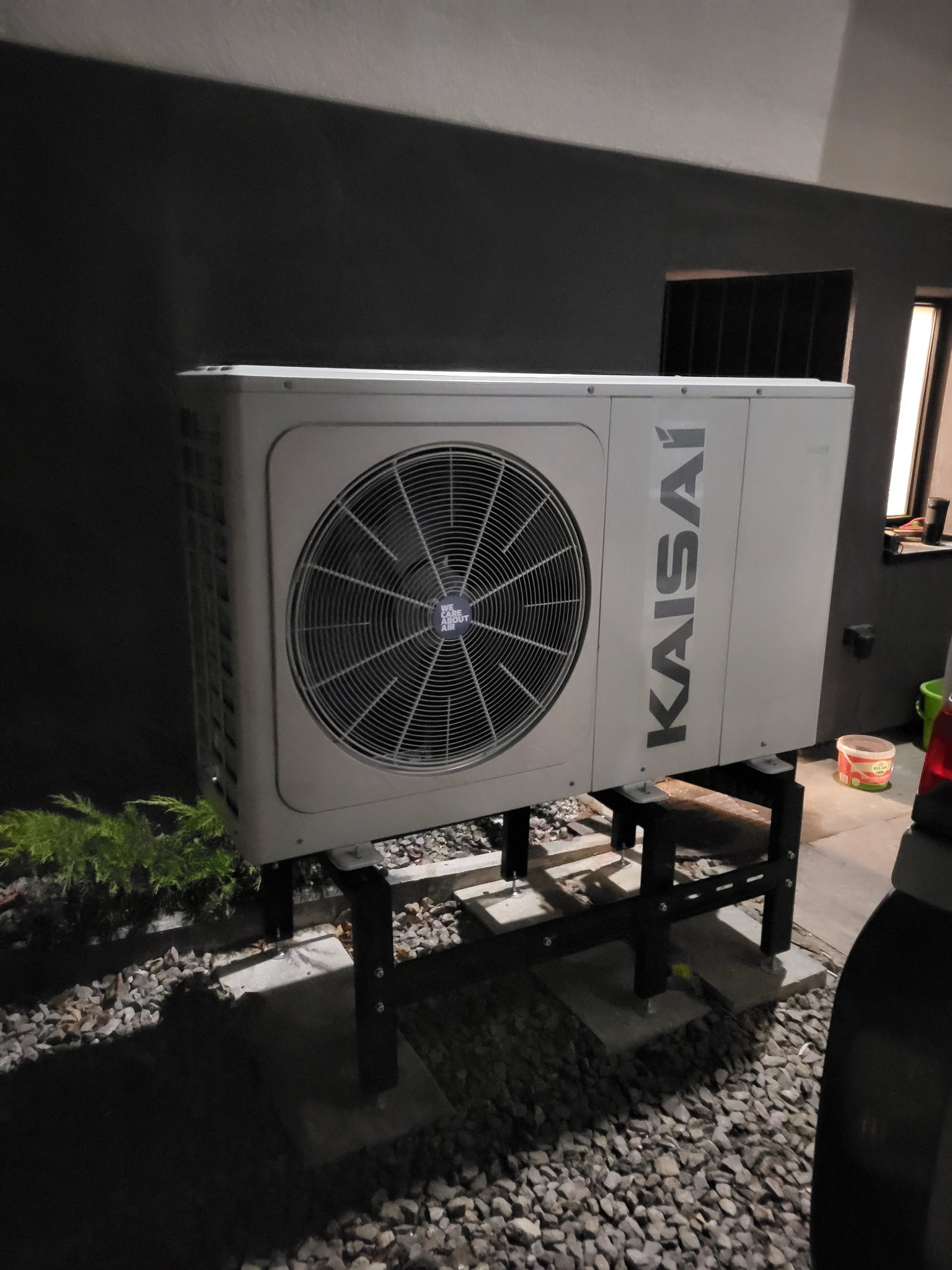 Zewnętrzna jednostka pompy ciepła marki Kaisai, zamontowana na podwyższeniu z metalowych profili, na tle grafitowej ściany budynku i żwirowego podłoża.