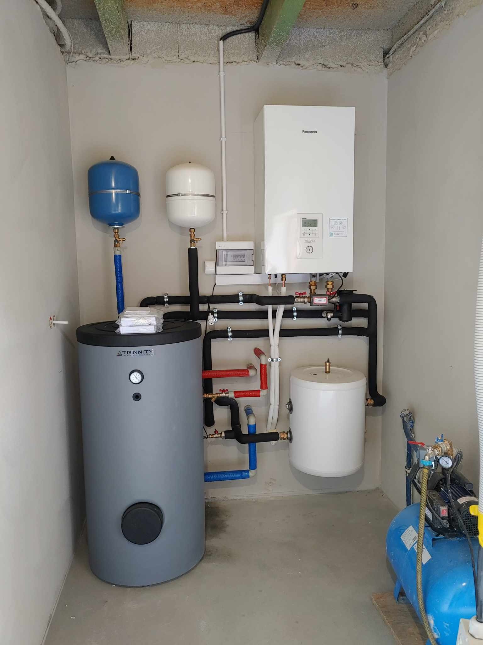 Kompletna instalacja grzewcza w kotłowni: szary zbiornik akumulacyjny Trinity, biały piec gazowy Panasonic, dwa zbiorniki przeponowe (niebieski i biały), liczne rury i zawory, oraz niebieski...