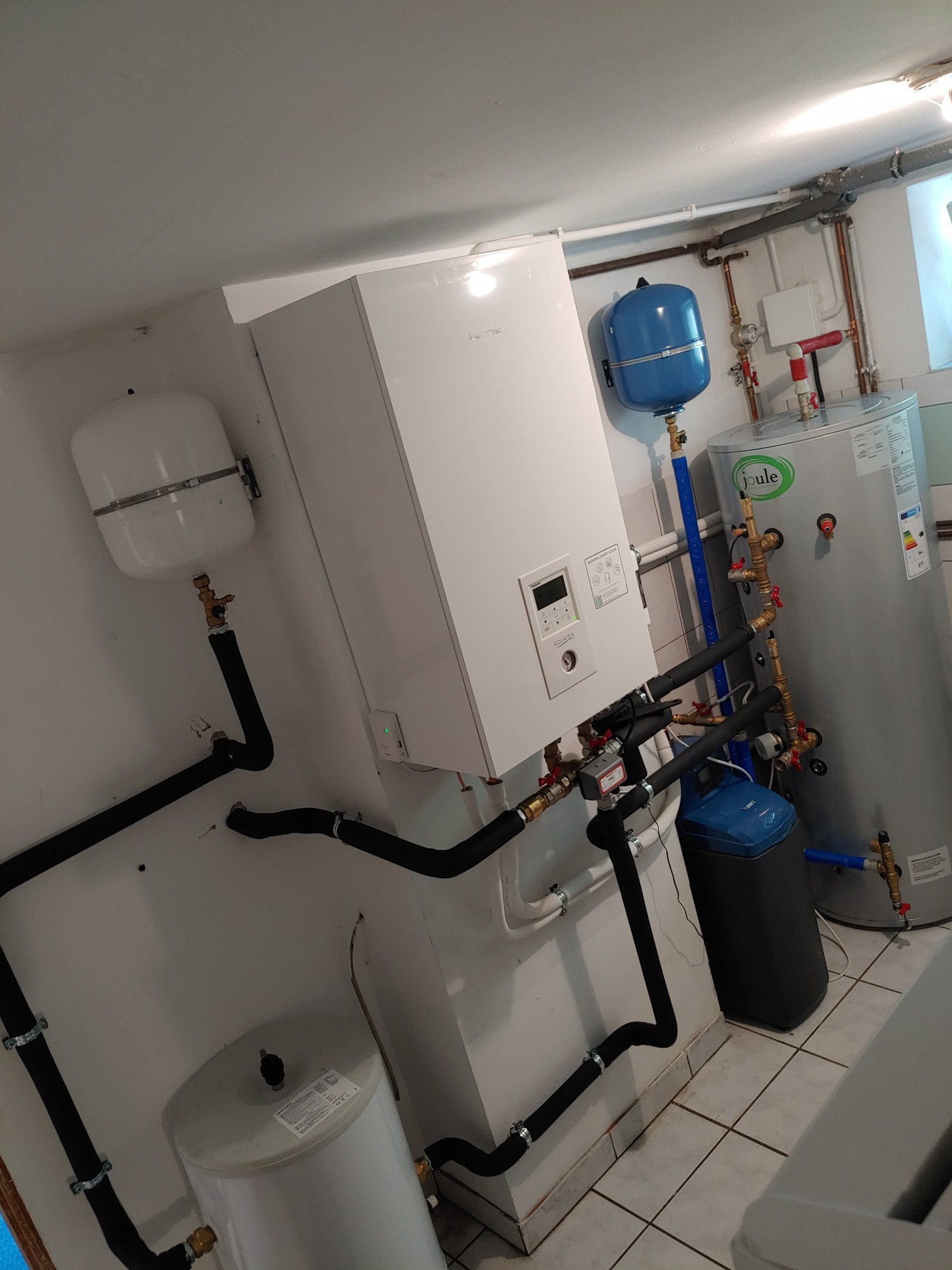 Zestaw urządzeń grzewczych w kotłowni: biały piec wiszący, zbiorniki wyrównawcze, zasobnik ciepłej wody Joule i filtr wody, połączone czarnymi rurami w izolacji, na tle białych ścian i płytek...