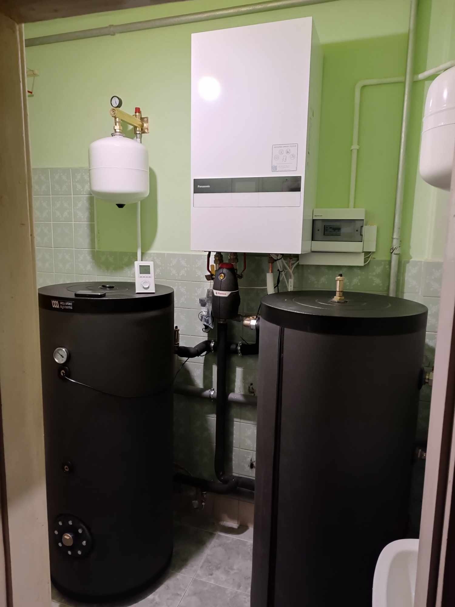 Instalacja z dwoma czarnymi zbiornikami akumulacyjnymi Aquatec Systems, białym zbiornikiem wyrównawczym z manometrem, białym piecem grzewczym Panasonic i skrzynką elektryczną na tle zielonej ściany...