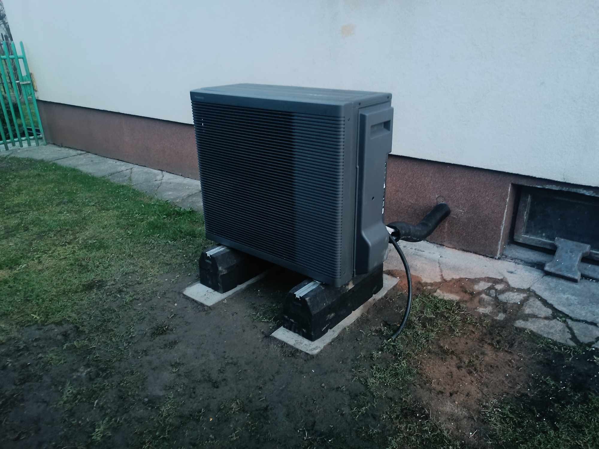 Zewnętrzna jednostka pompy ciepła Panasonic Aquarea, umieszczona na betonowych podstawach w pobliżu ściany budynku z widocznym oknem piwnicznym i rurą odprowadzającą skropliny, otoczona trawnikiem...