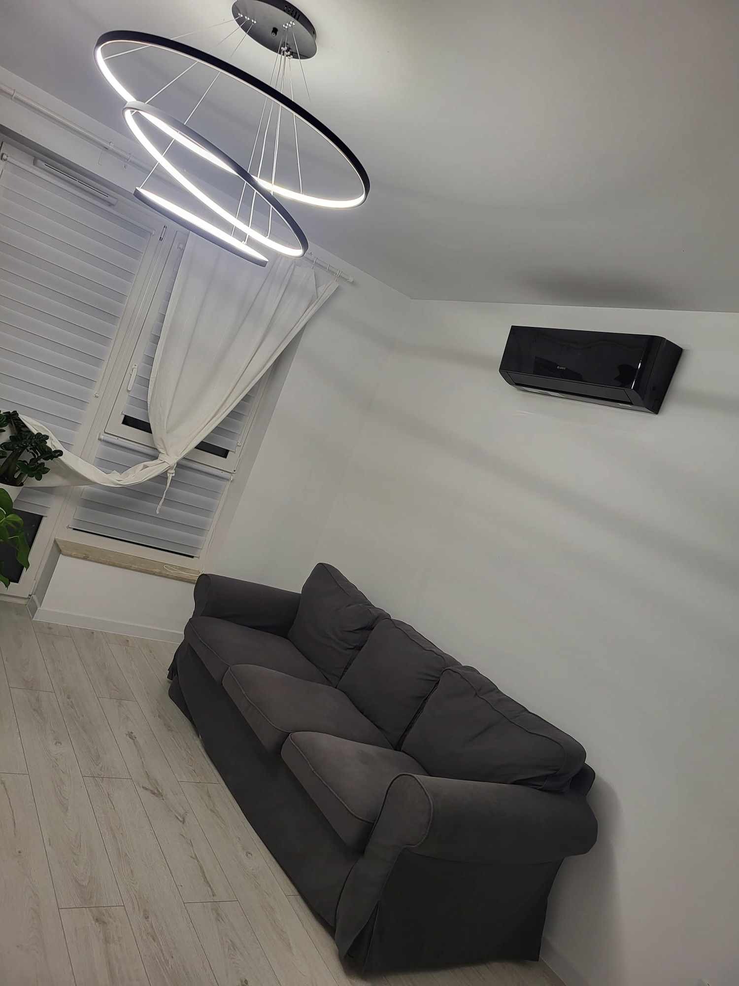 Czarny klimatyzator ścienny w nowoczesnym salonie z szarą sofą, pod trzema okrągłymi lampami LED i oknem z białymi zasłonami.