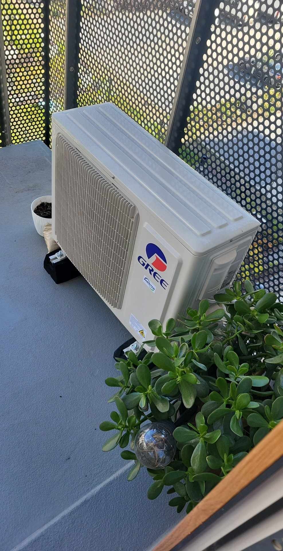Klimatyzator marki Gree zamontowany na balkonie w Krakowie, widoczny przez ażurową balustradę, obok doniczka z kwiatami.