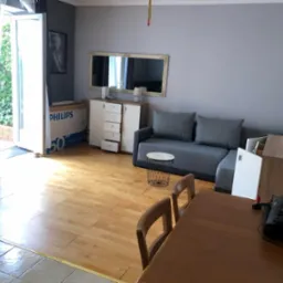 Widok na salon z drewnianą podłogą, szarą sofą, stolikiem kawowym z metalową podstawą, otwartym balkonem i widocznym kartonem po telewizorze Philips.