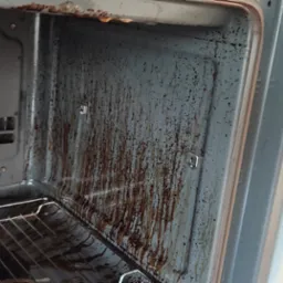 Wnętrze brudnego piekarnika z przypalonym tłuszczem i resztkami jedzenia na ściankach, kratce i wentylatorze, widok 'przed' czyszczeniem.